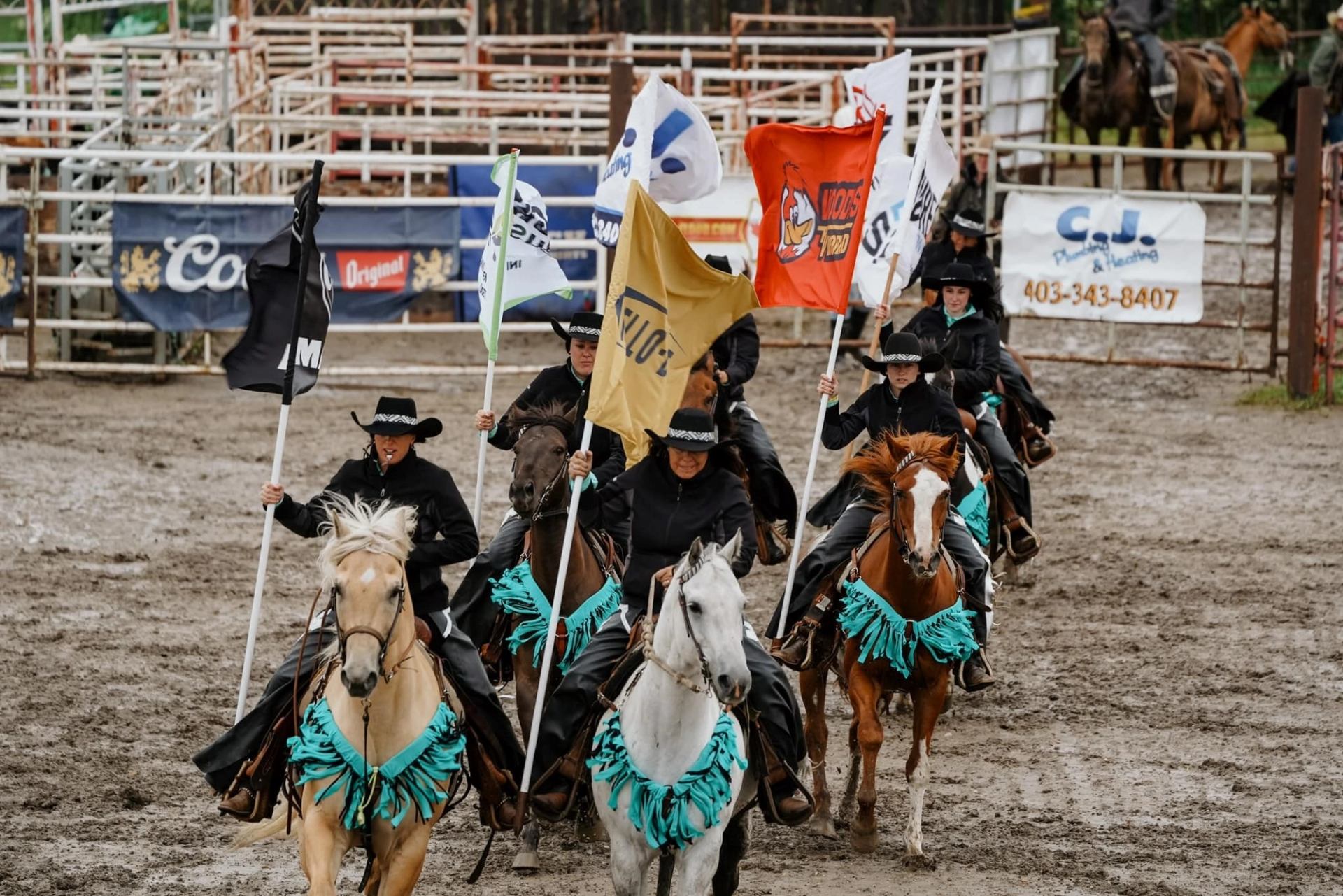 Mounted riders carrying colorful flags into a muddy rodeo arena during the Daines Ranch Pro Rodeo.