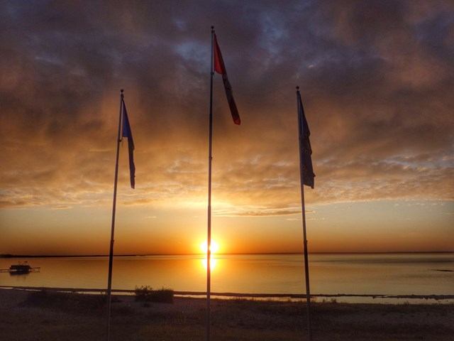 Three flagpoles with flags silhouetted against sunset over calm lake and clouds.