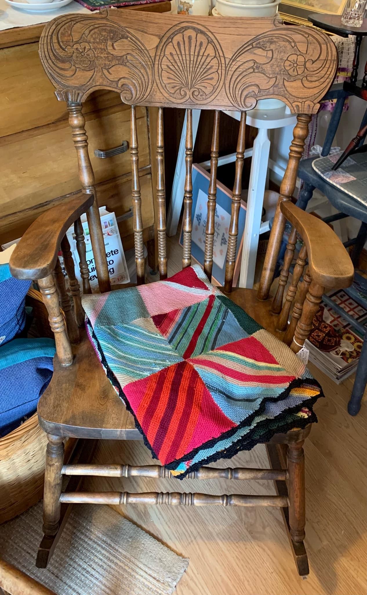 Wooden rocking chair with intricate carvings and a colorful knitted quilt draped over the seat.