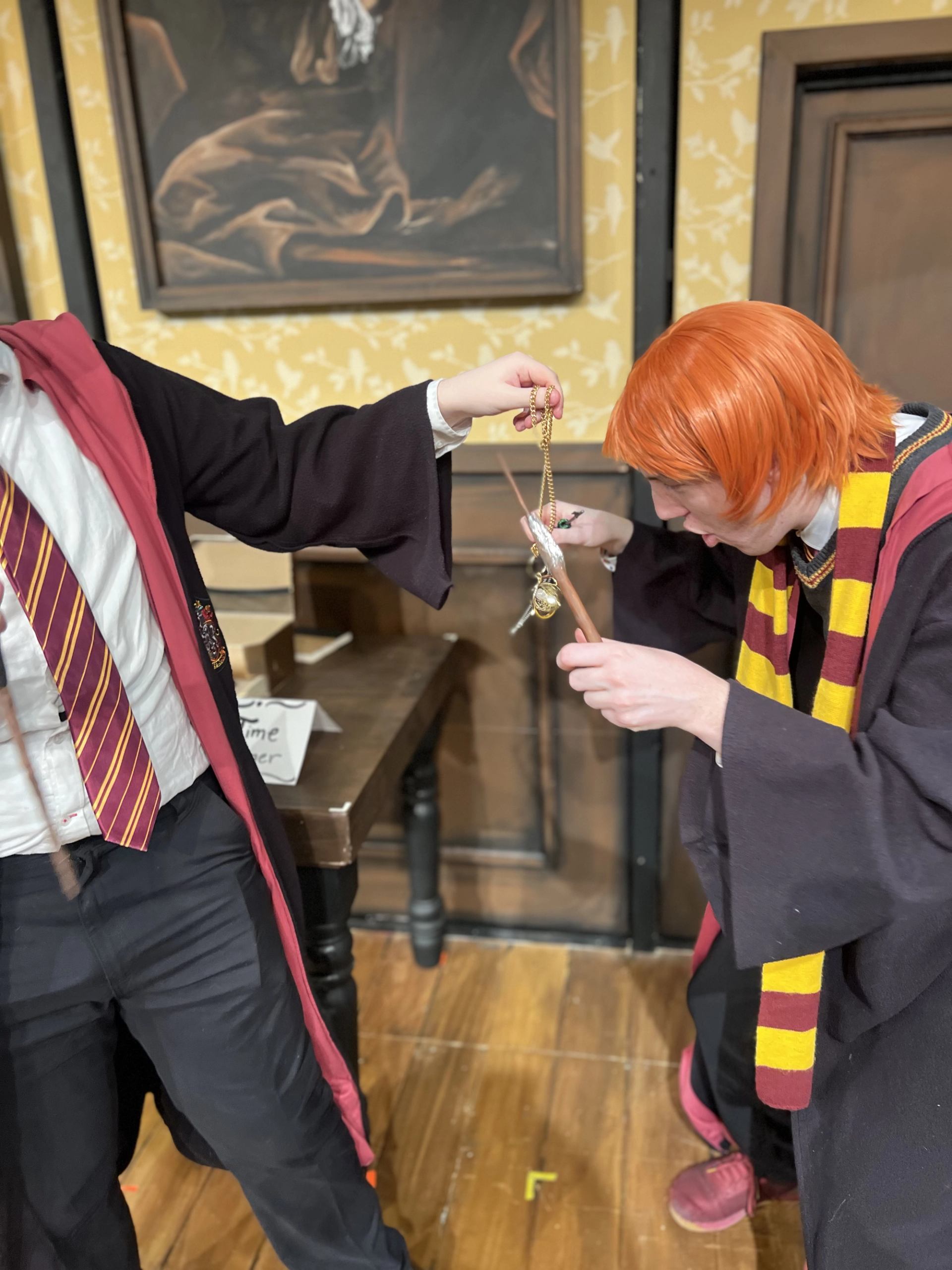 Two people in wizard costumes examine a golden necklace prop in a decorated room.