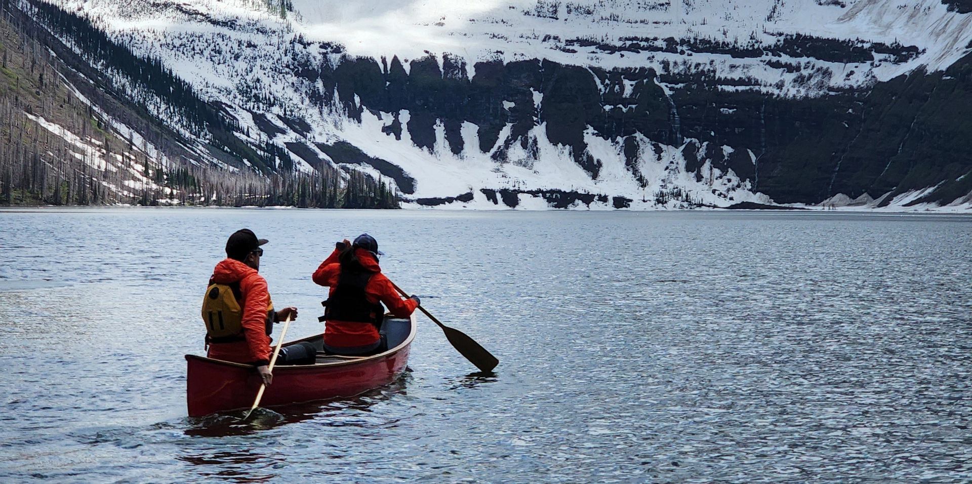 Two canoers paddle on a lake with mountains in the background.