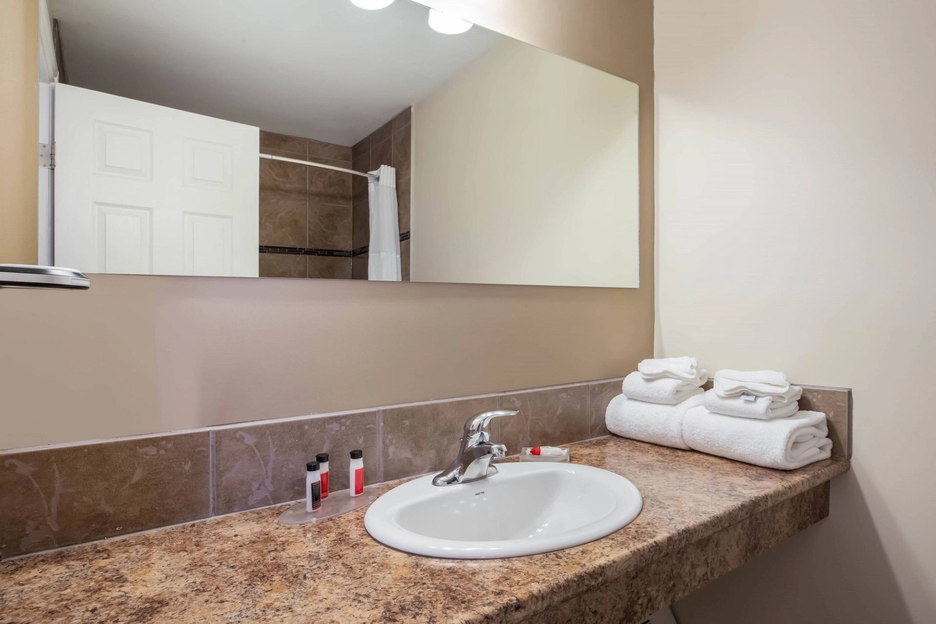 A bathroom countertop with a white sink, stacked towels, and small toiletries, with a shower curtain visible in the background.