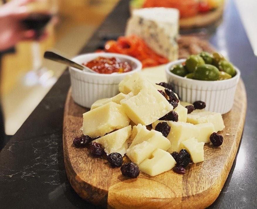 Wooden board topped with sliced Italian cheeses, olives, dried fruit, and small bowls of spreads.