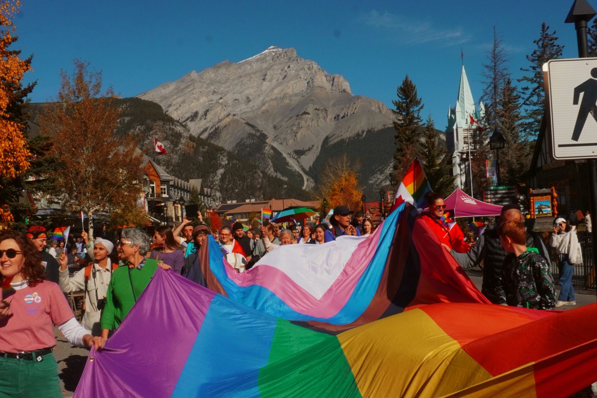 People hold a large rainbow flag at a pride parade, with mountains, tents, and fall trees behind.