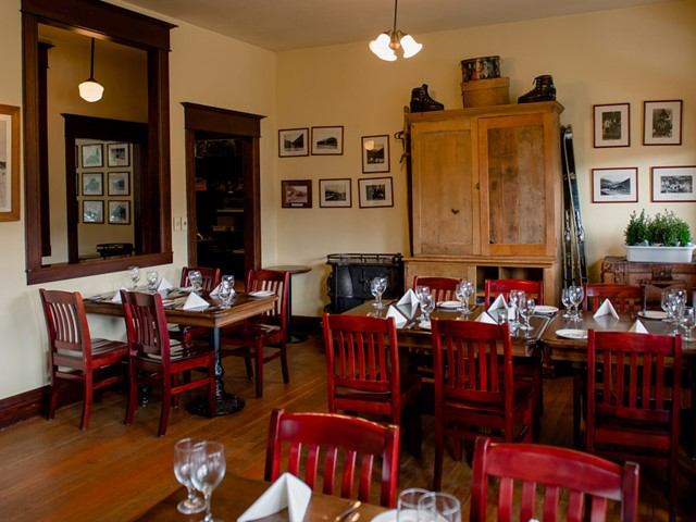 Cozy dining room with red wooden chairs, set tables, and folded napkins. Walls adorned with framed photos, a wooden cabinet, and warm lighting.