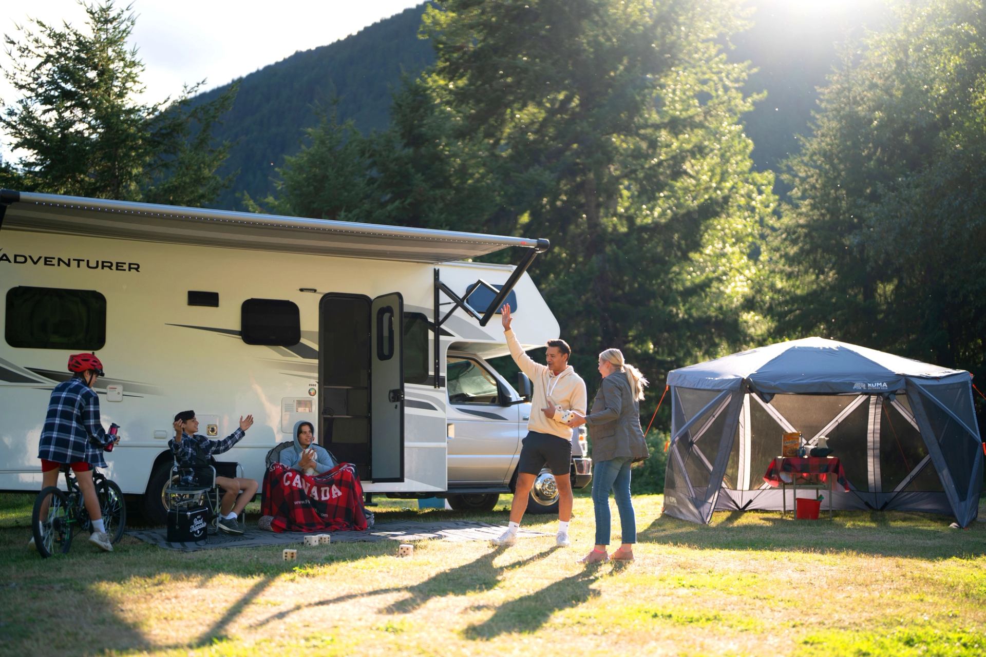 Group camping near RV with trees and mountains.