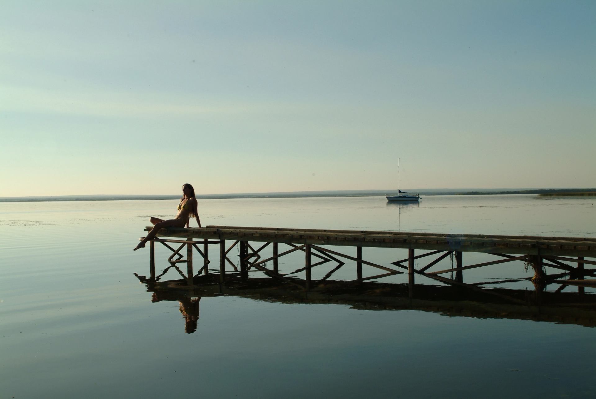 Person sitting on wooden dock over calm water, sailboat in distance.