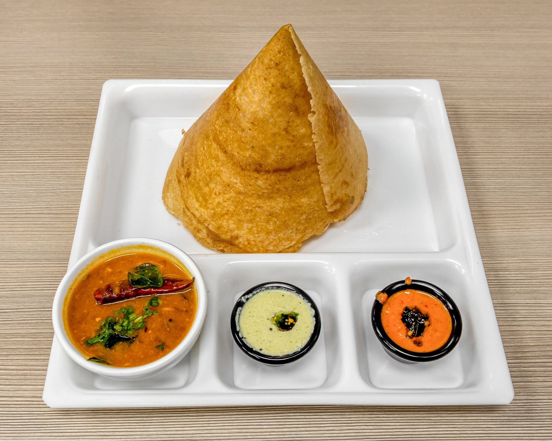 Cone-shaped dosa served with sambar and two chutneys on a white tray.