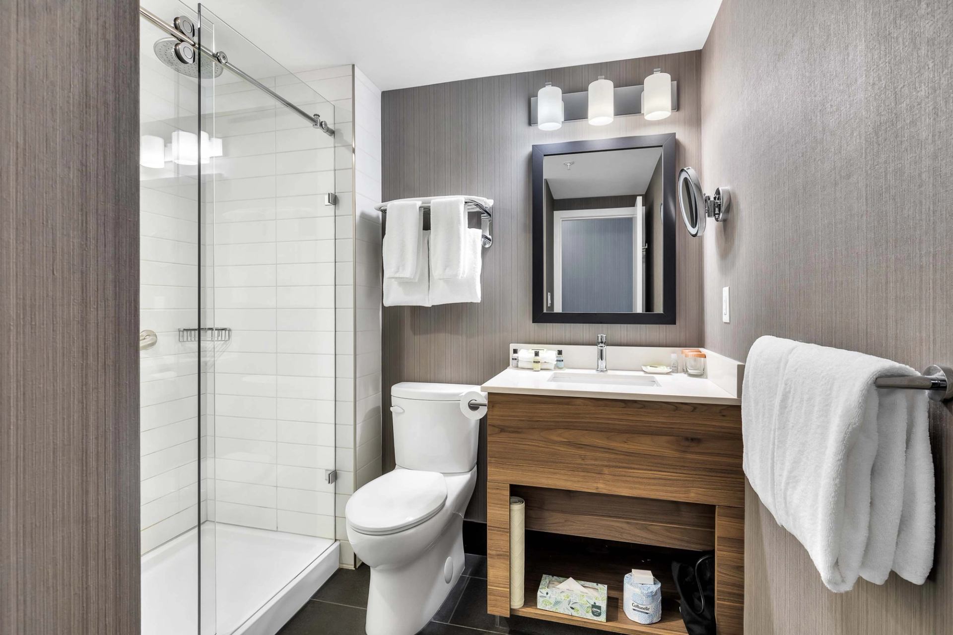 Modern bathroom with glass shower, toilet, wood vanity, mirror and white towels.