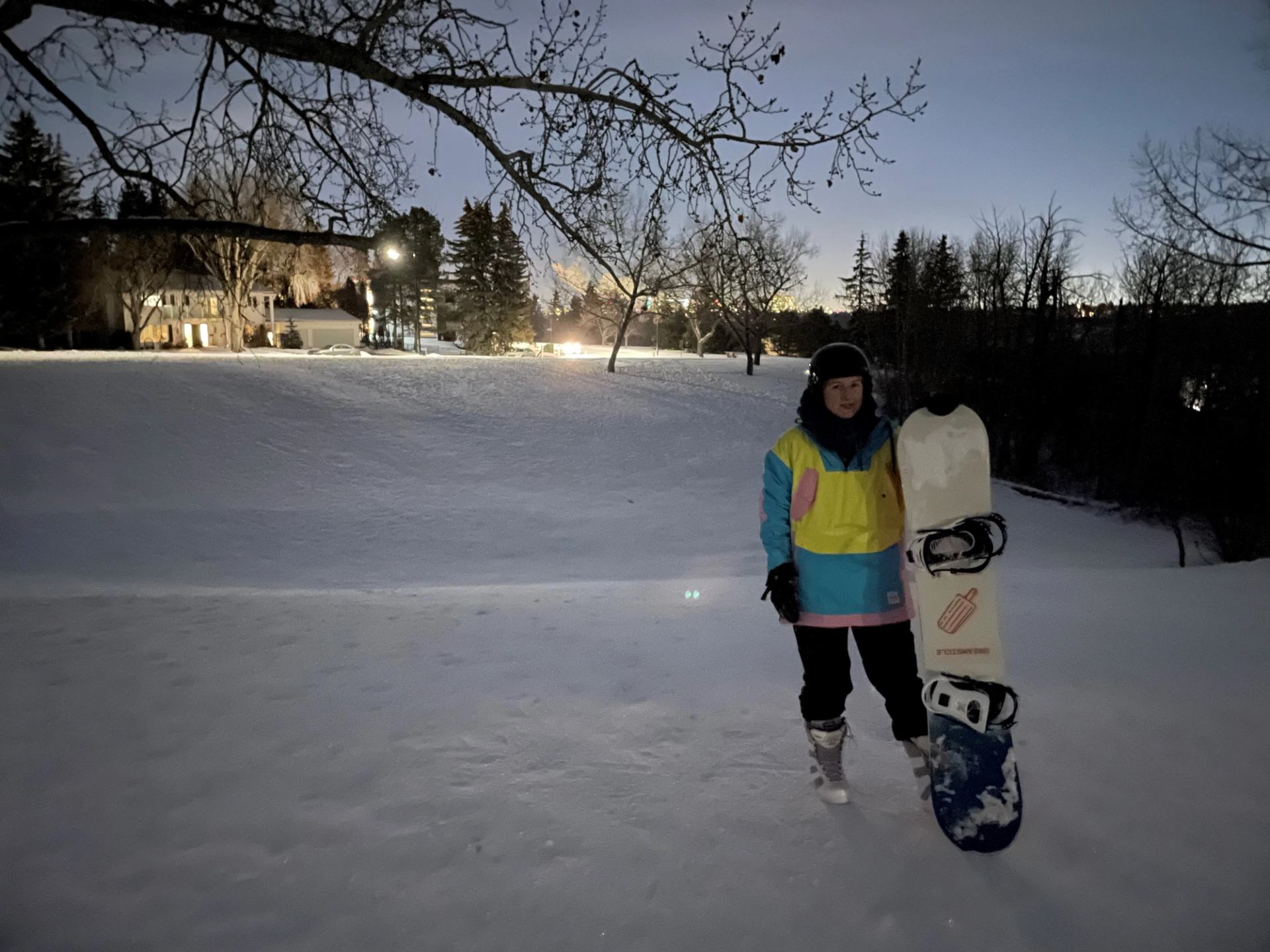 Snow-covered field with a snowboarder in winter gear holding a snowboard near trees and buildings.