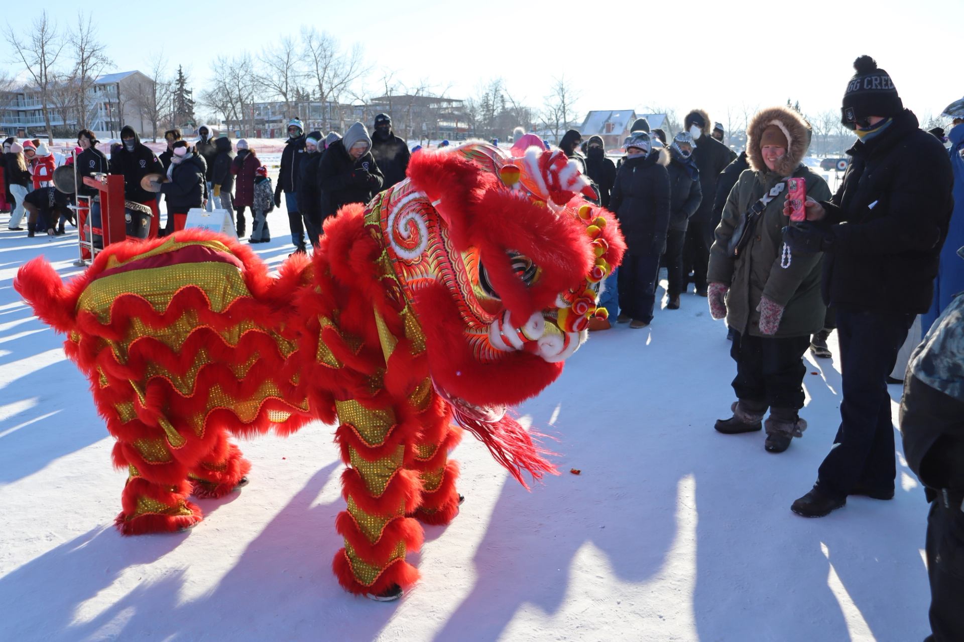 Red and gold lion dance costumes on ice path with spectators walking in background.