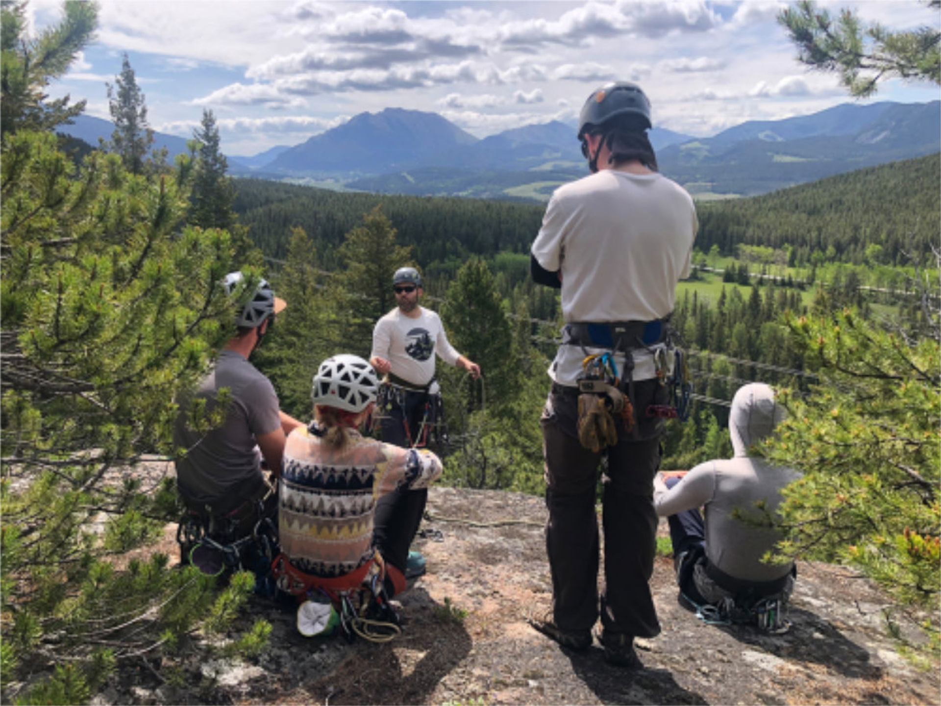 Rock climbers wearing helmets gather on a cliff ledge overlooking forested mountains.