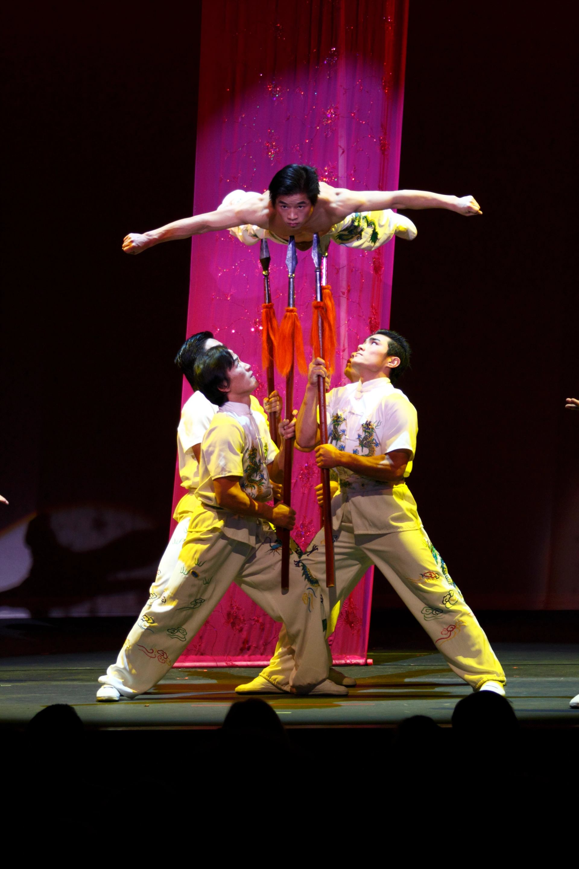 Acrobats balance performer on upright spears before pink curtain on dim stage.
