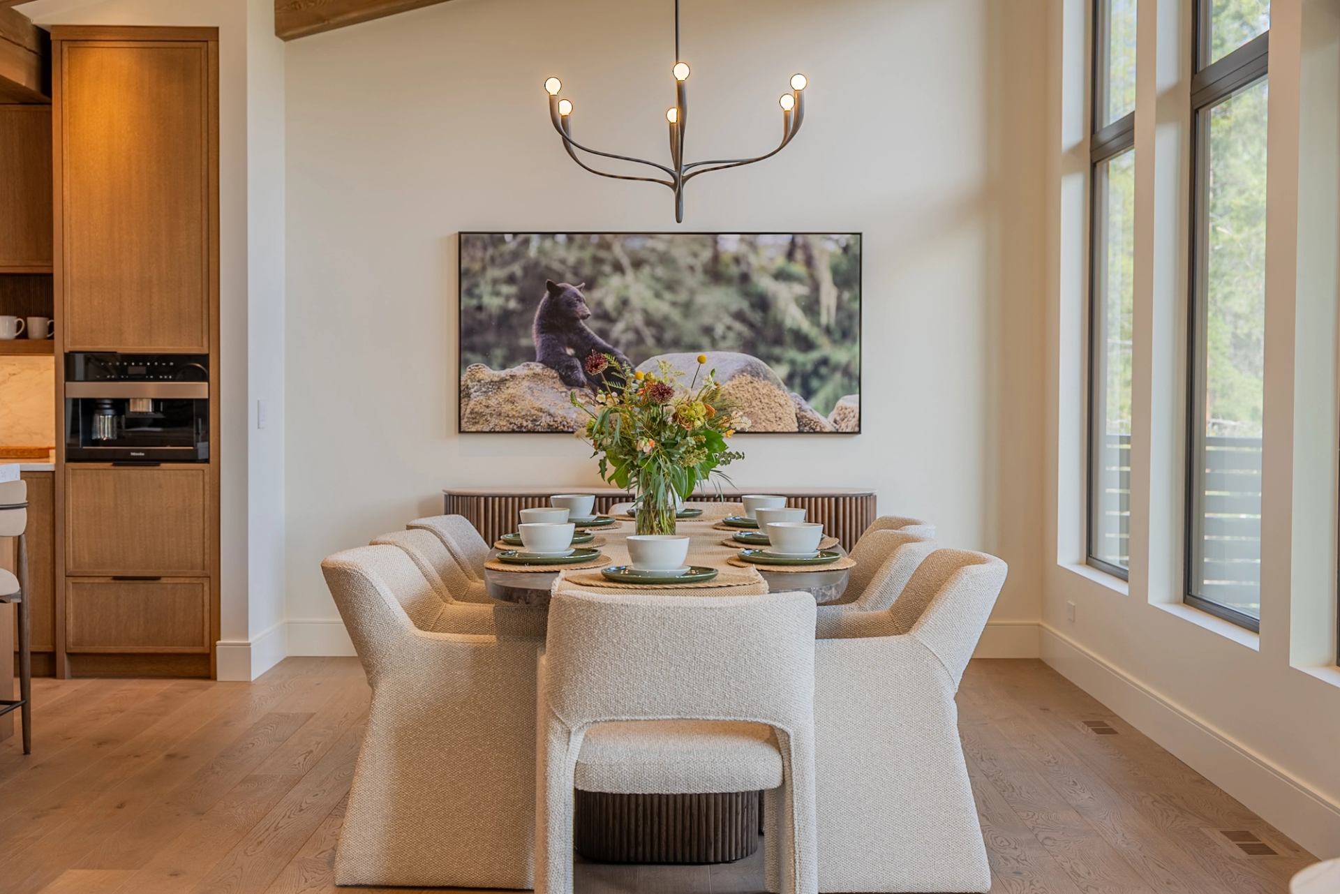 A beautifully set dining table with upholstered chairs sits beneath a modern chandelier, featuring a floral centerpiece and large windows that fill the room with natural light.