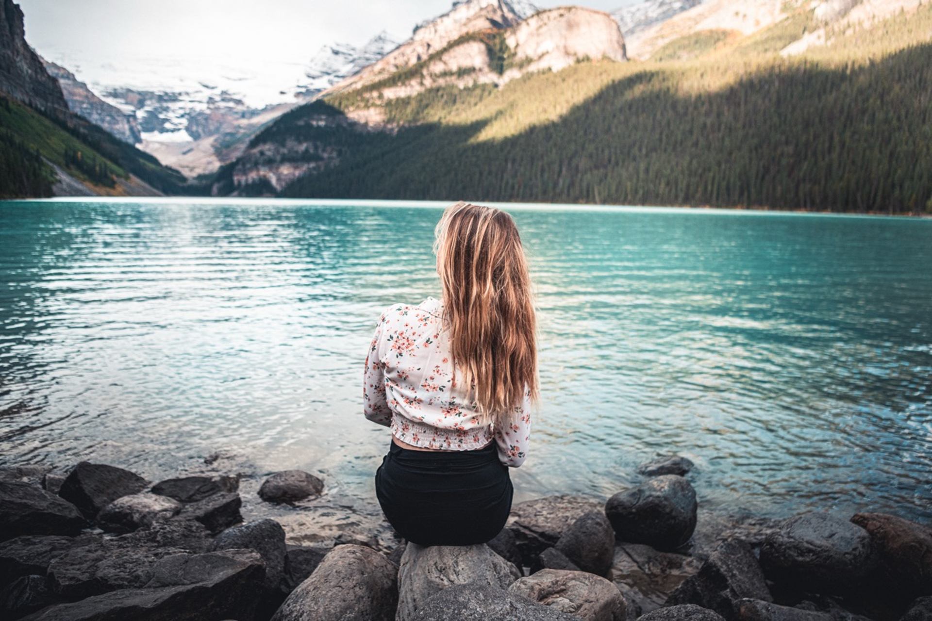 Person with long hair sits on lakeshore rocks facing turquoise water and mountains.