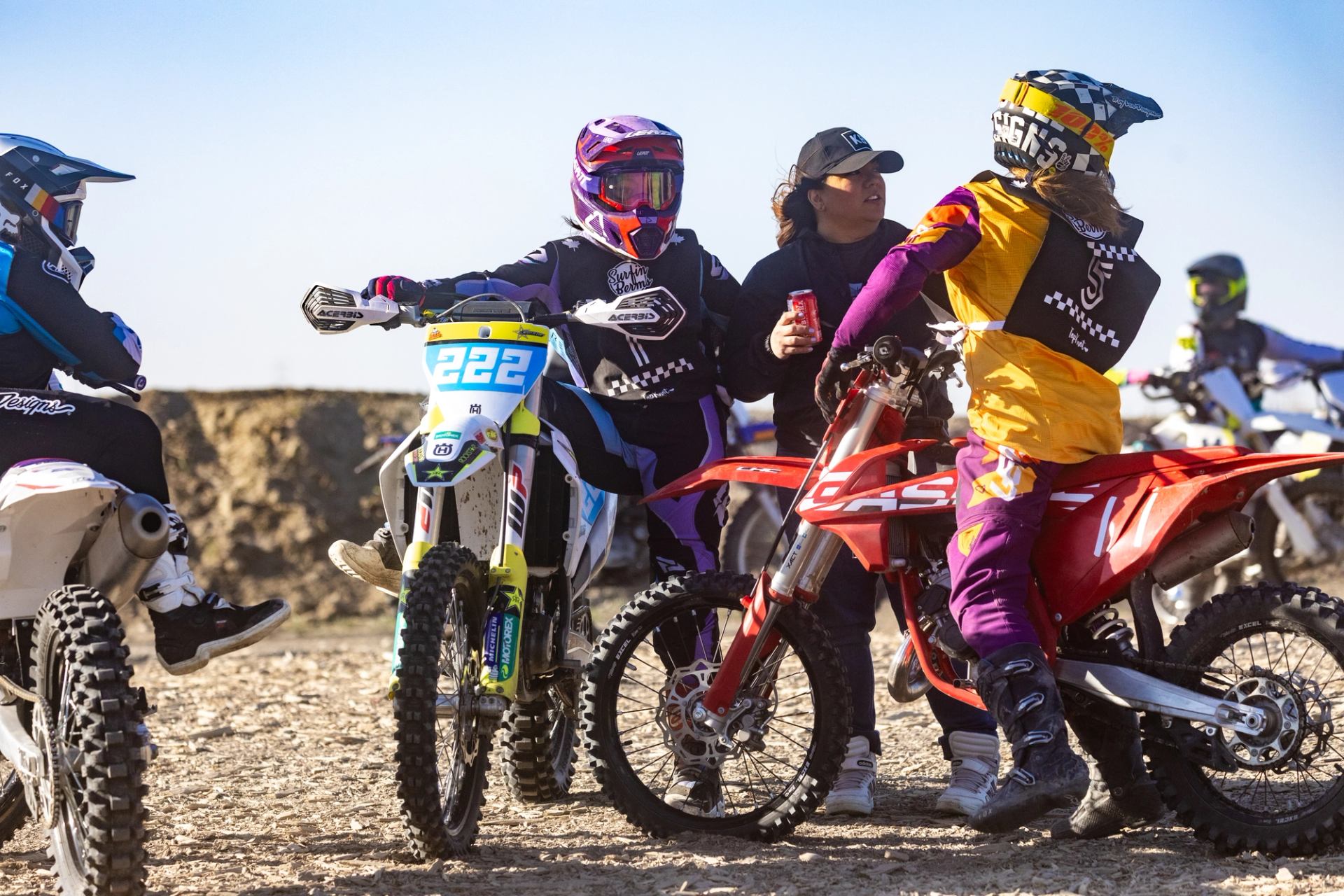 Motocross riders in gear gathered on dirt track under clear skies.
