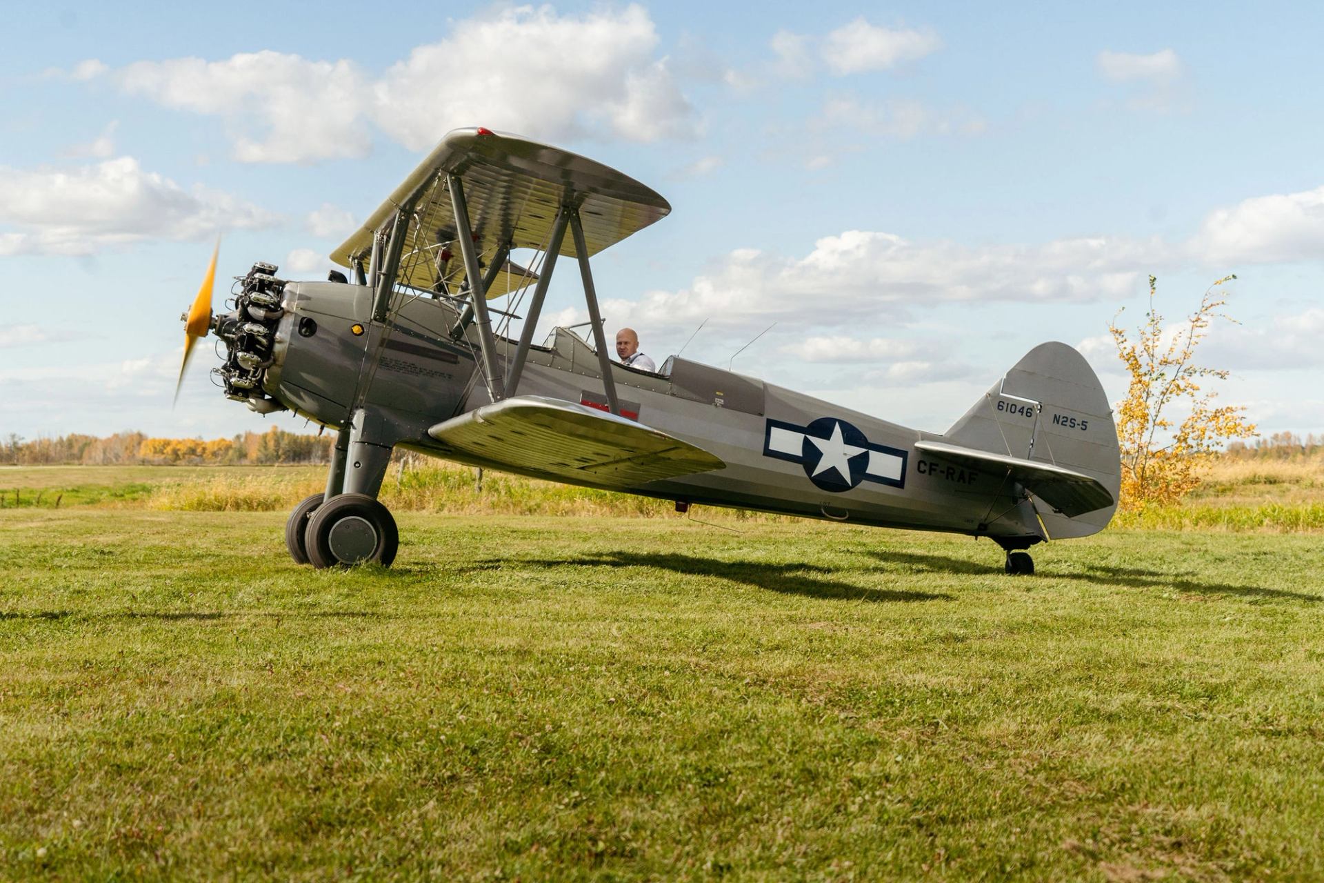 Vintage biplane with spinning propeller sits on grassy airfield under partly cloudy sky.