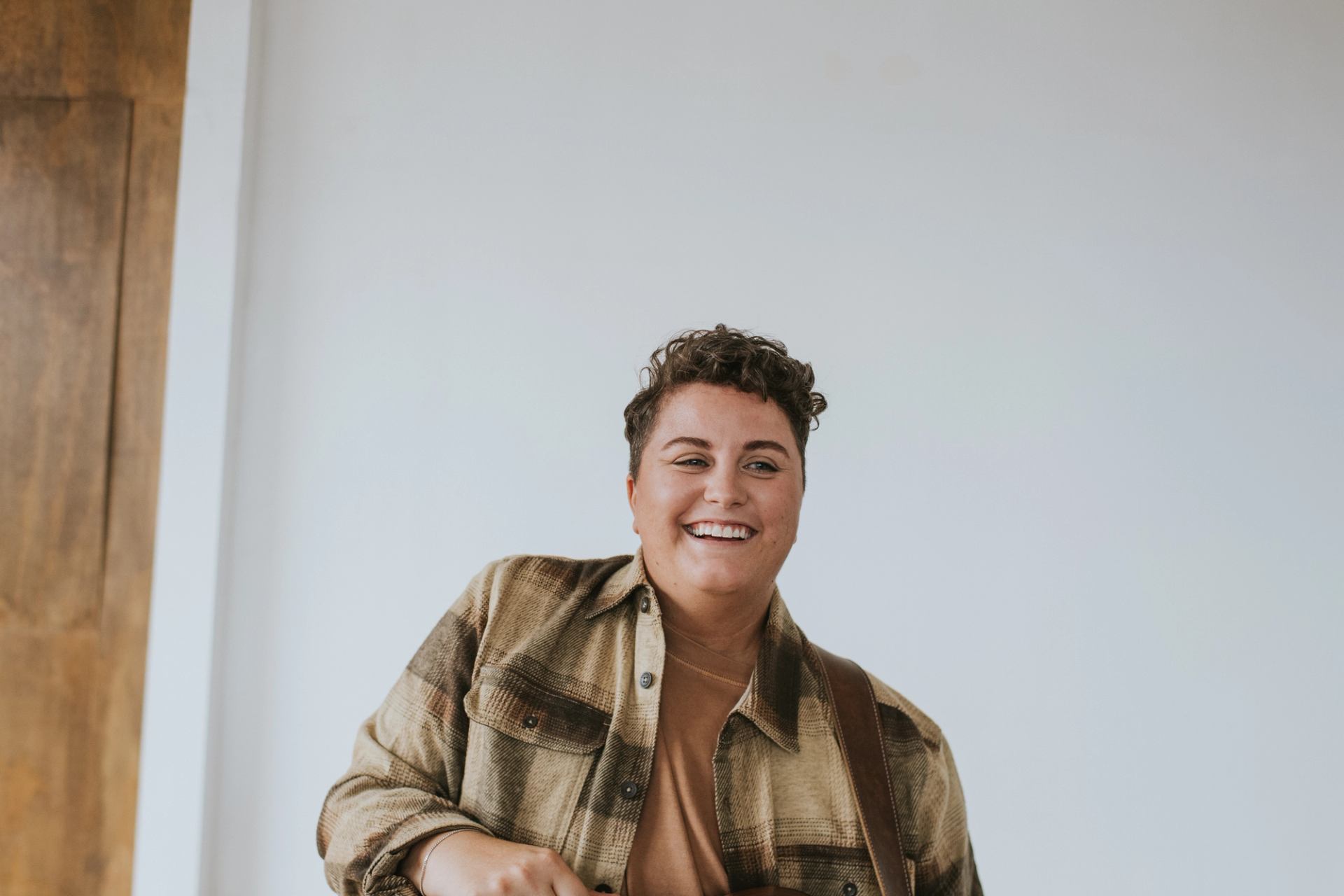 Person in plaid shirt stands indoors against plain white wall, looking toward camera.