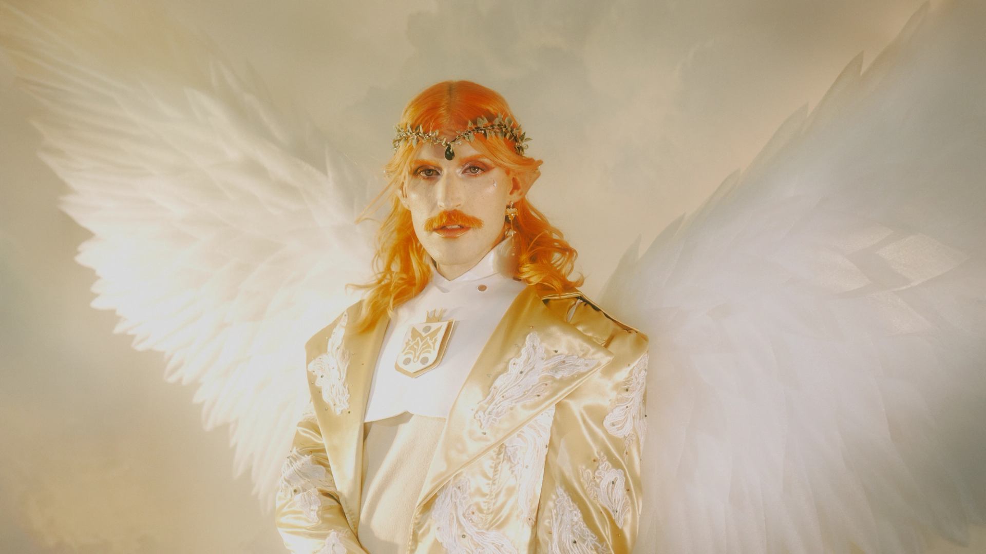 Person in ornate gold suit and crown stands with large white feathered wings.