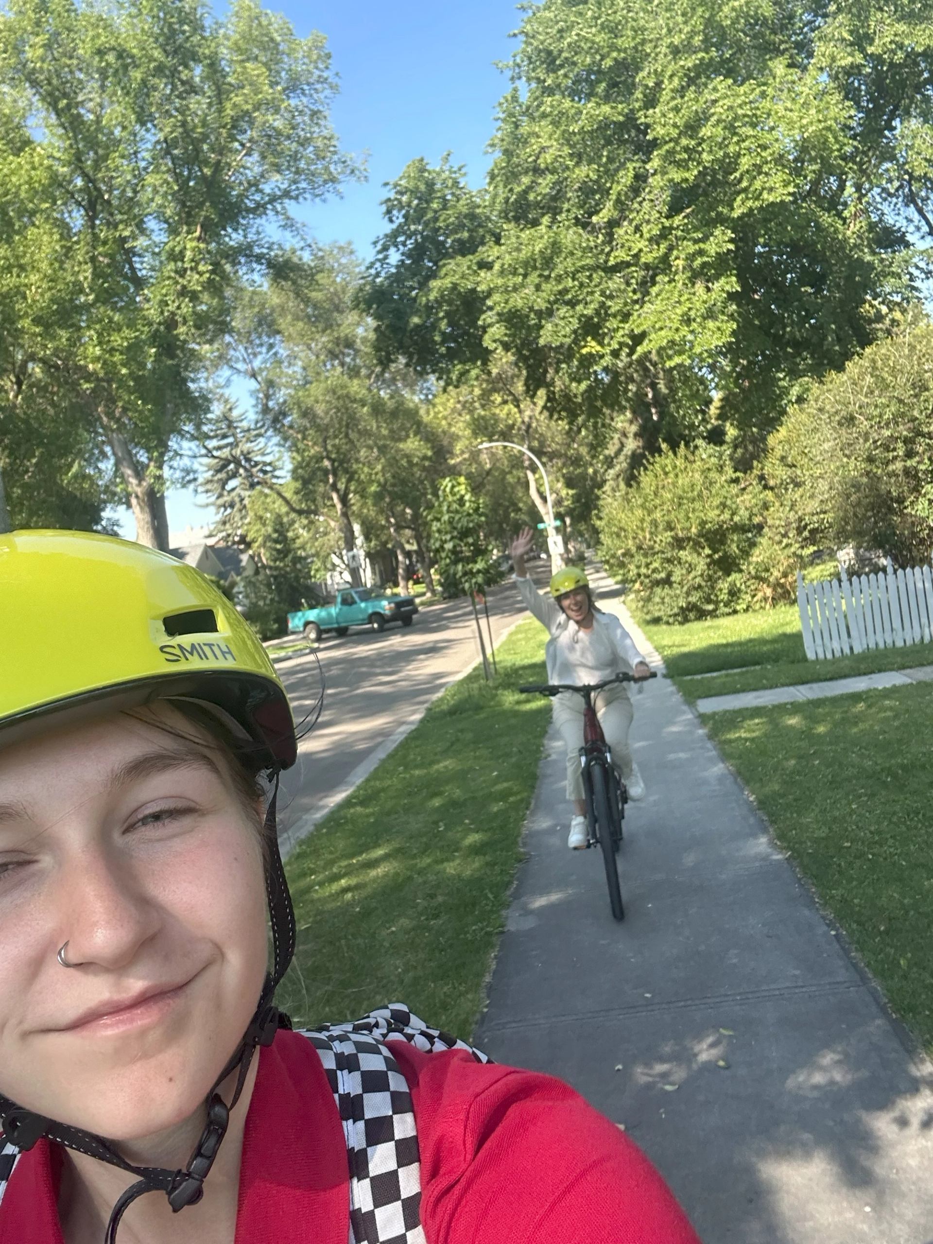 Two cyclists ride through a sunny park path with trees and grass on either side.