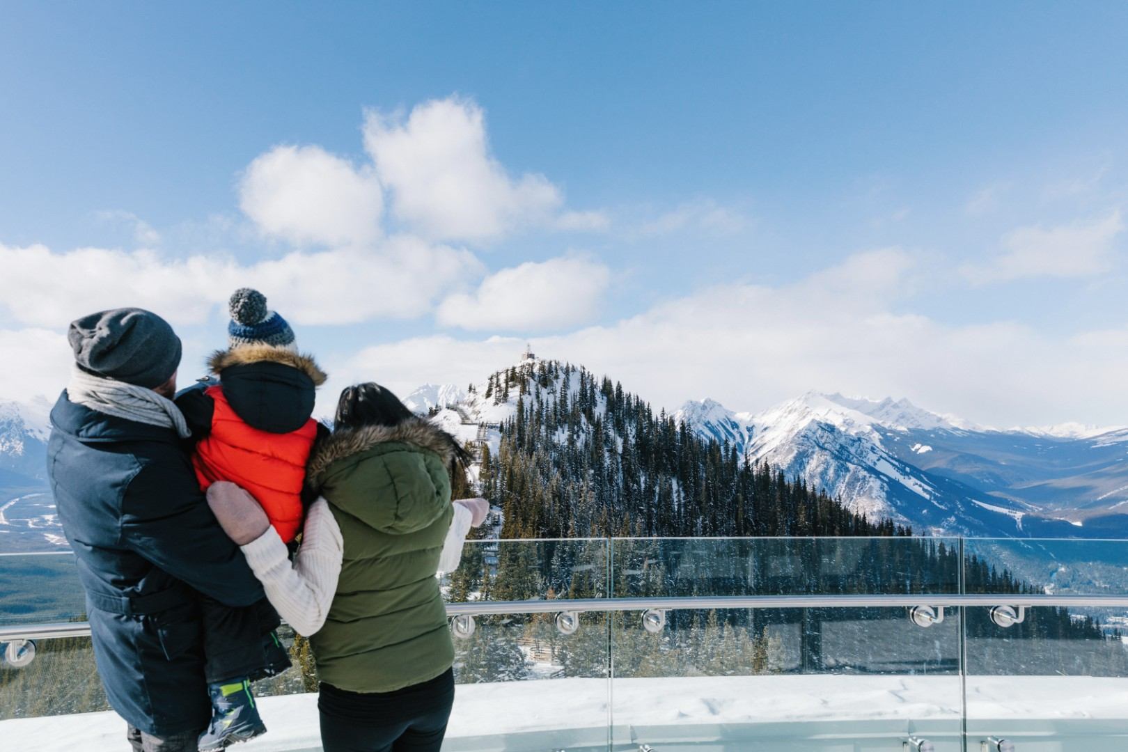 Three bundled people on a glass lookout deck facing snowy mountain peaks and forest.