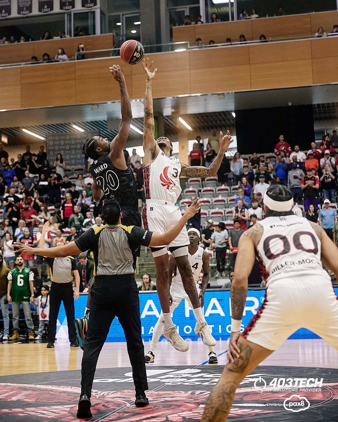 Basketball player shoots ball during game, spectators and players visible in indoor arena.