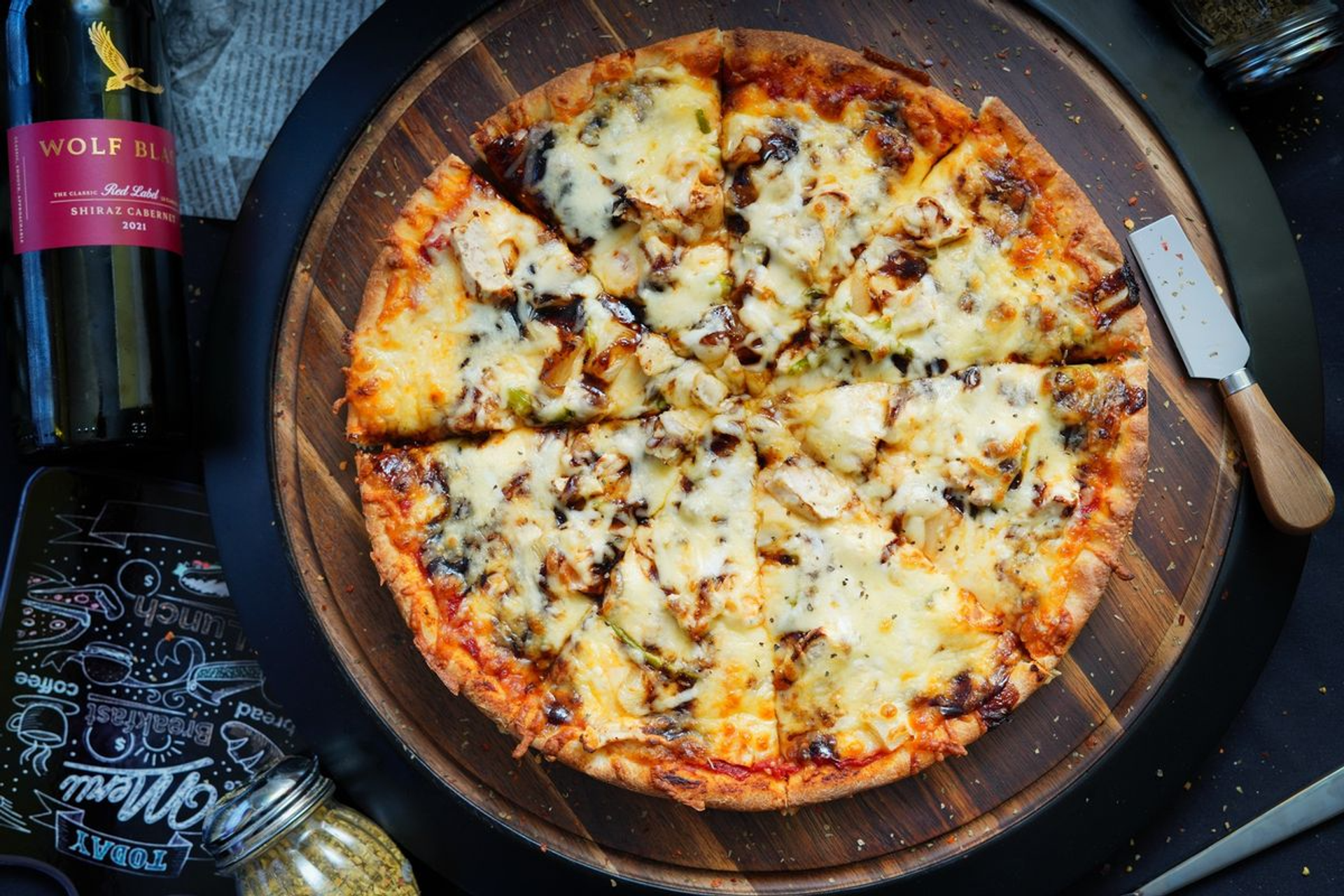 Sliced cheese and chicken pizza on a wooden board beside a wine bottle.