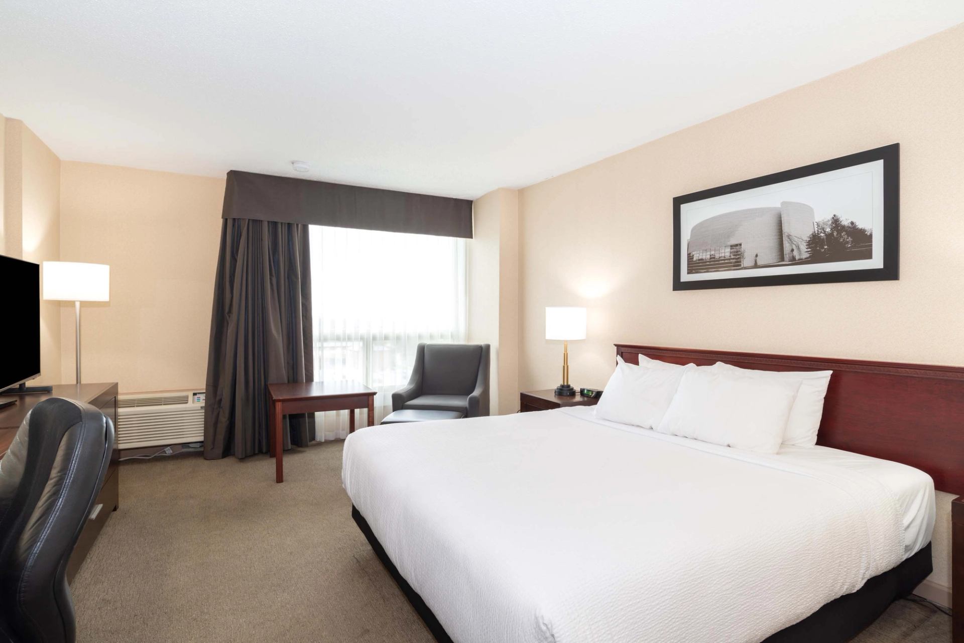 A hotel room with a king bed, desk, armchair, and large window.
