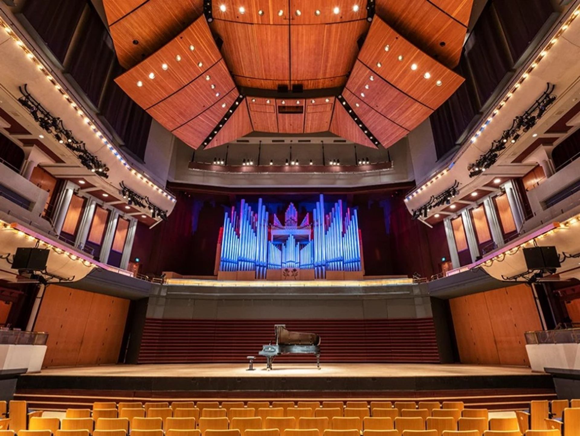Concert hall interior with grand piano on stage and illuminated pipe organ backdrop.
