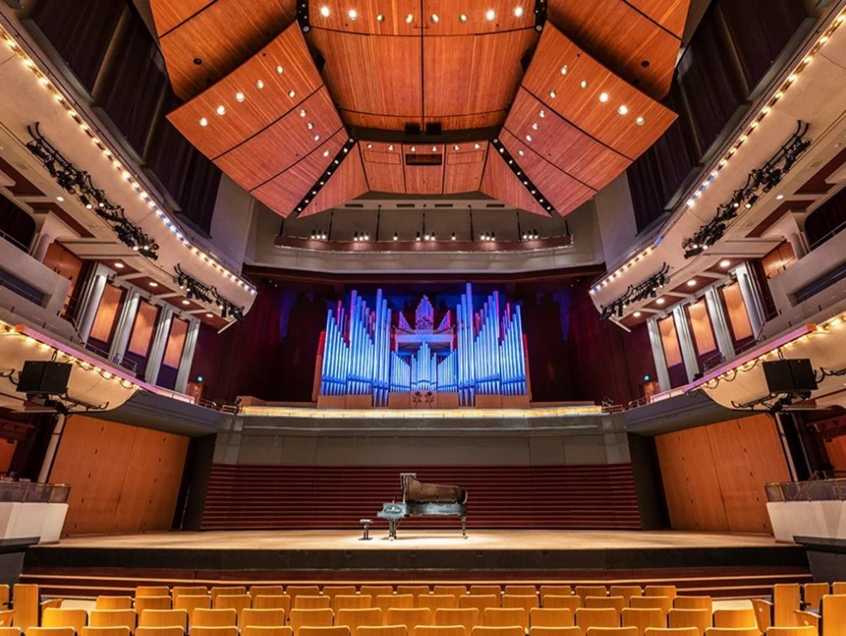 Concert hall interior with grand piano on stage and illuminated pipe organ backdrop.