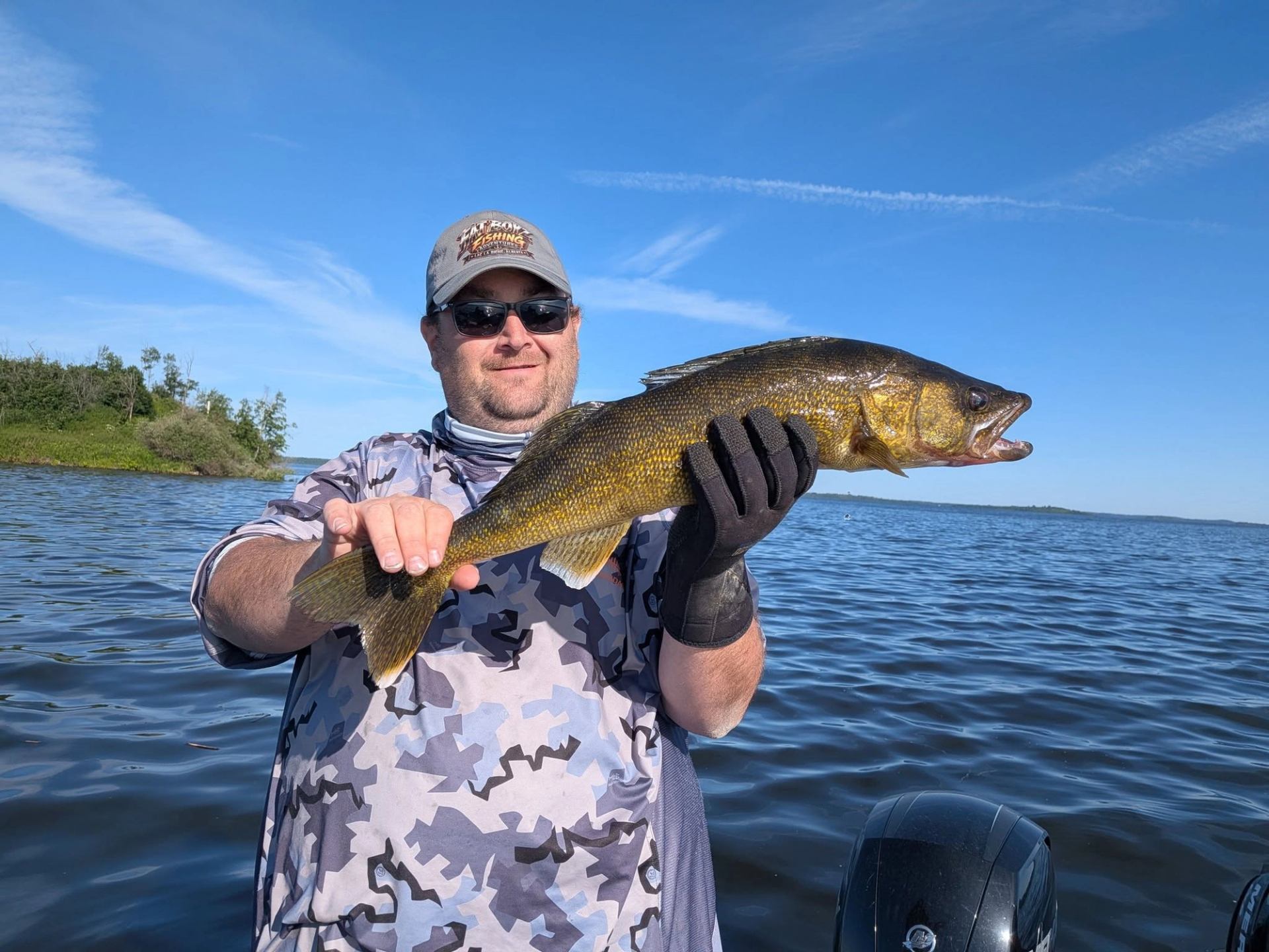 Angler on boat holding large walleye over calm lake with distant shoreline.