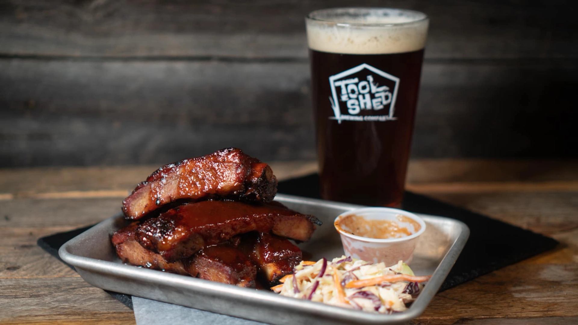 Tray of sauced pork ribs with coleslaw and dip beside a Tool Shed beer.
