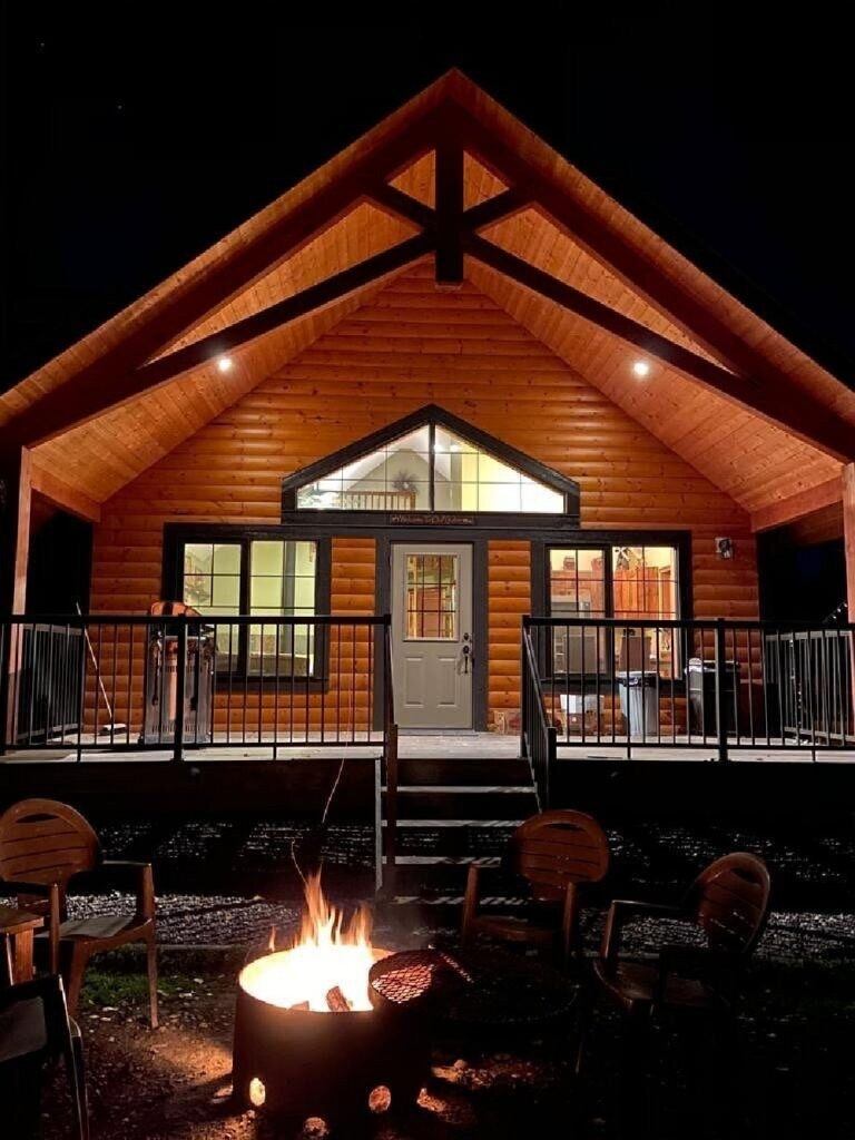 Lit cabin at night with fire pit and porch in a cozy forest setting.