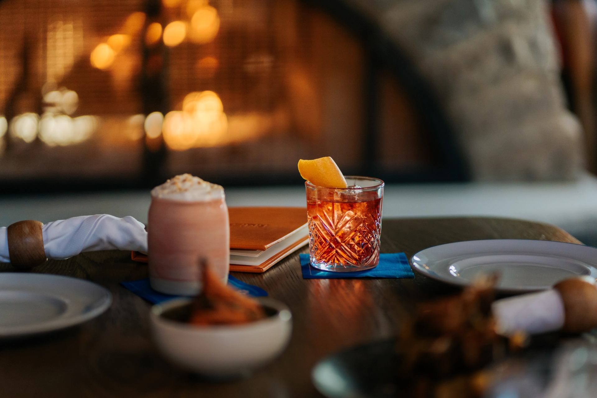 A table set with two drinks, including a cocktail with an orange slice, and plates,  glowing fireplace.