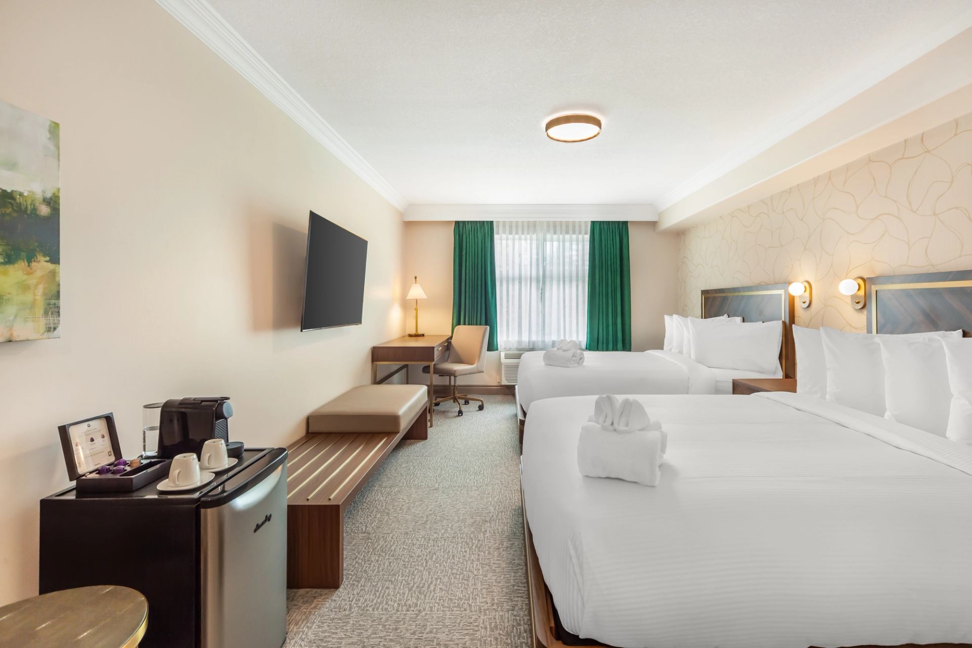 Spacious hotel room with two large beds, crisp white linens, a wall-mounted TV, desk, and mini-fridge.