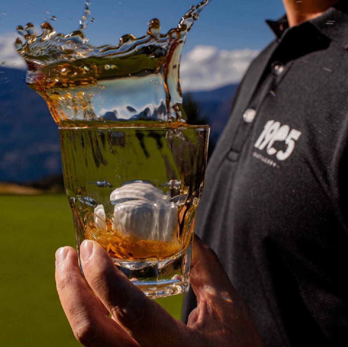 A man holding a glass of 1905 spirit with a golf ball splashed in it.