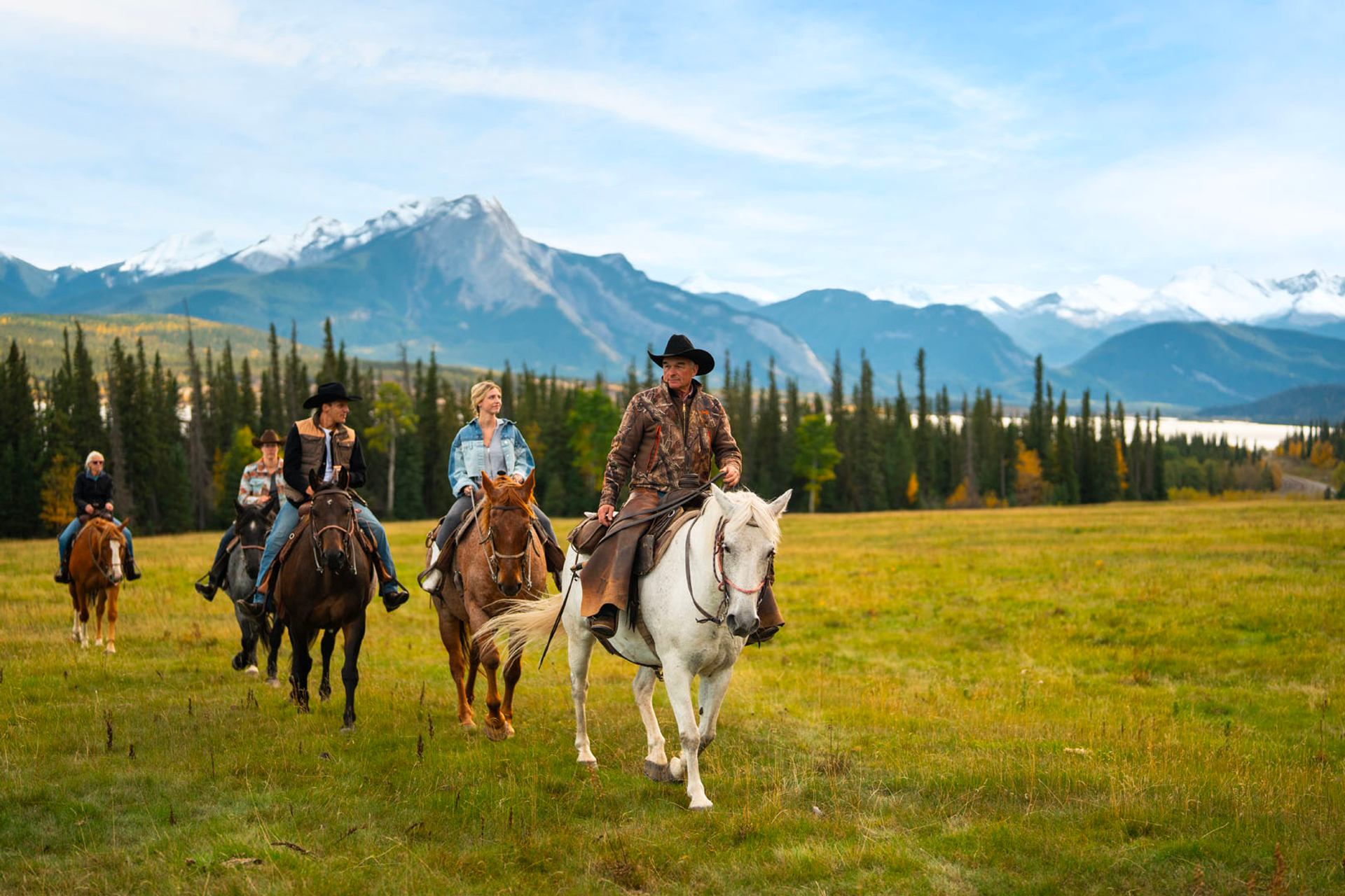 A group riding horses across a grassy meadow with tall mountains behind.