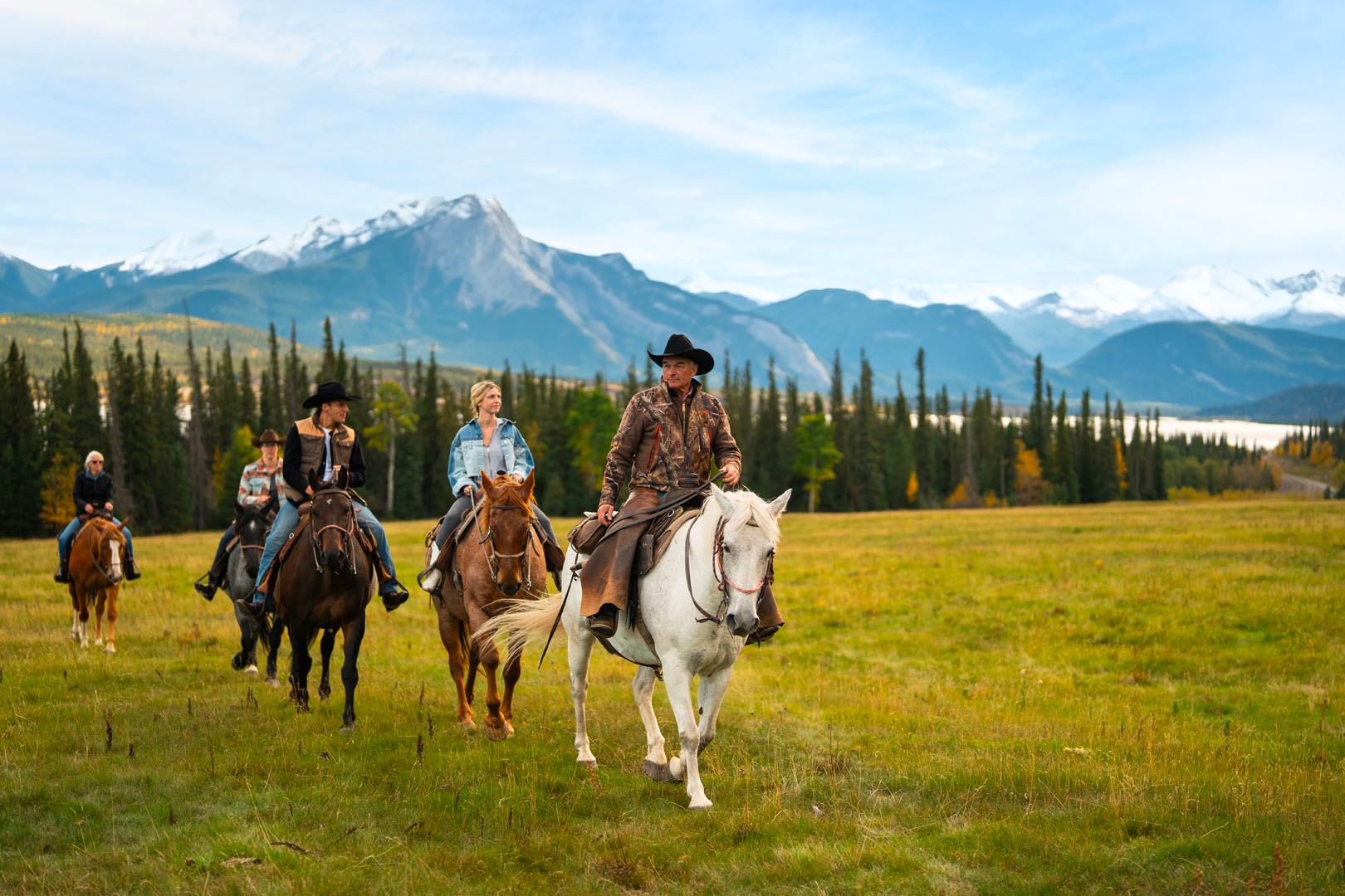 A group riding horses across a grassy meadow with tall mountains behind.