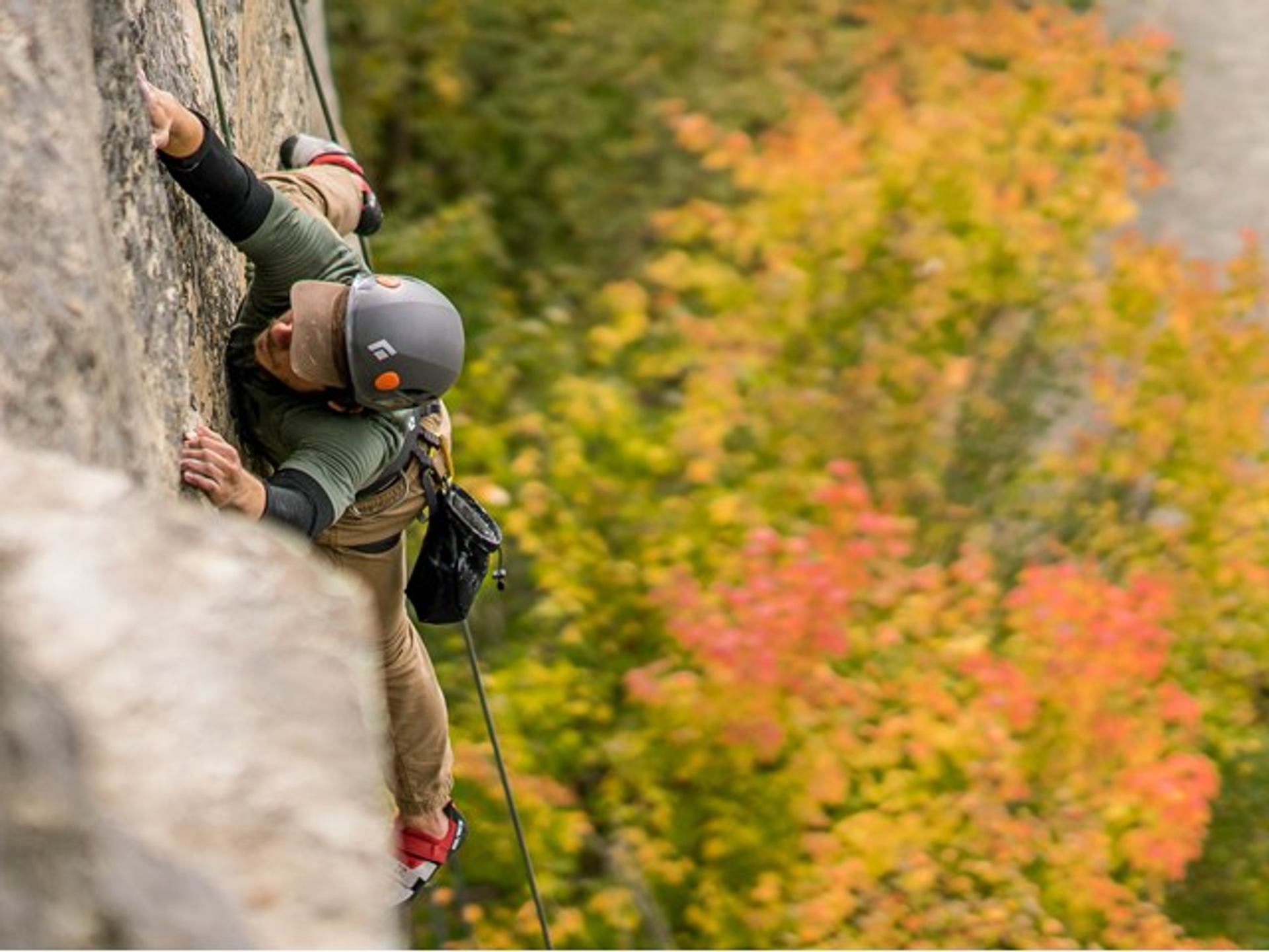 Guided climber ascending a vertical rock face with safety rope, surrounded by colorful autumn forest.