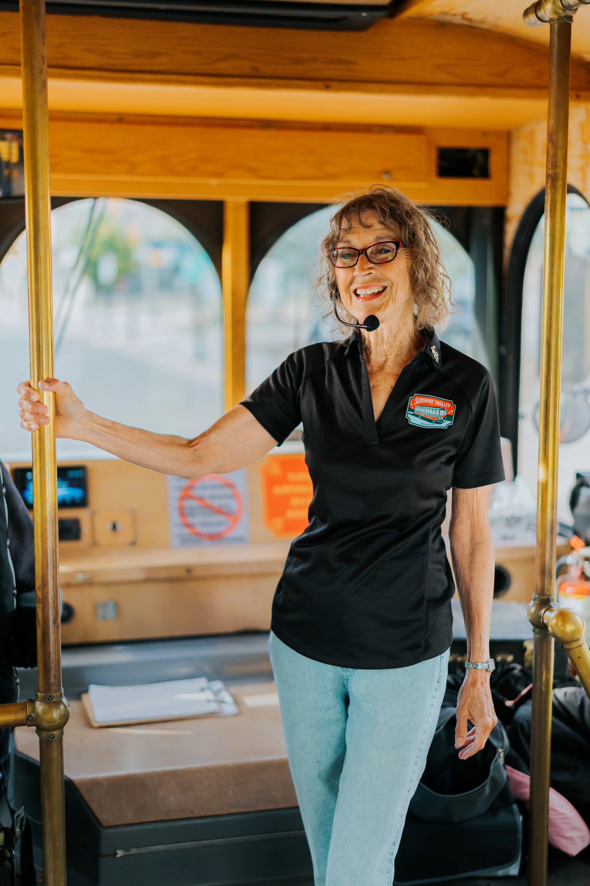 Tour guide wearing headset stands inside trolley bus, holding brass pole near driver area.