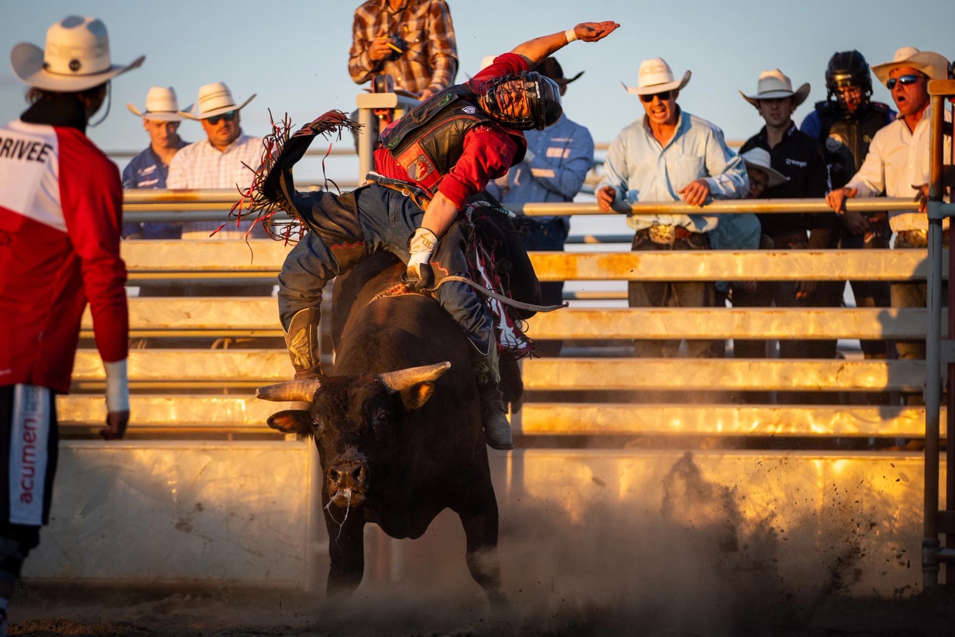 Bull rider grips bucking bull in dusty rodeo arena as spectators watch from stands.