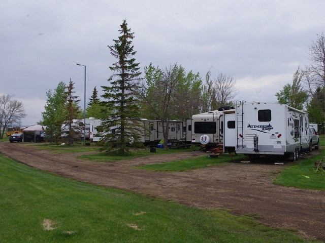 RVs parked along a dirt road at a campground with green grass, trees, and an overcast sky.