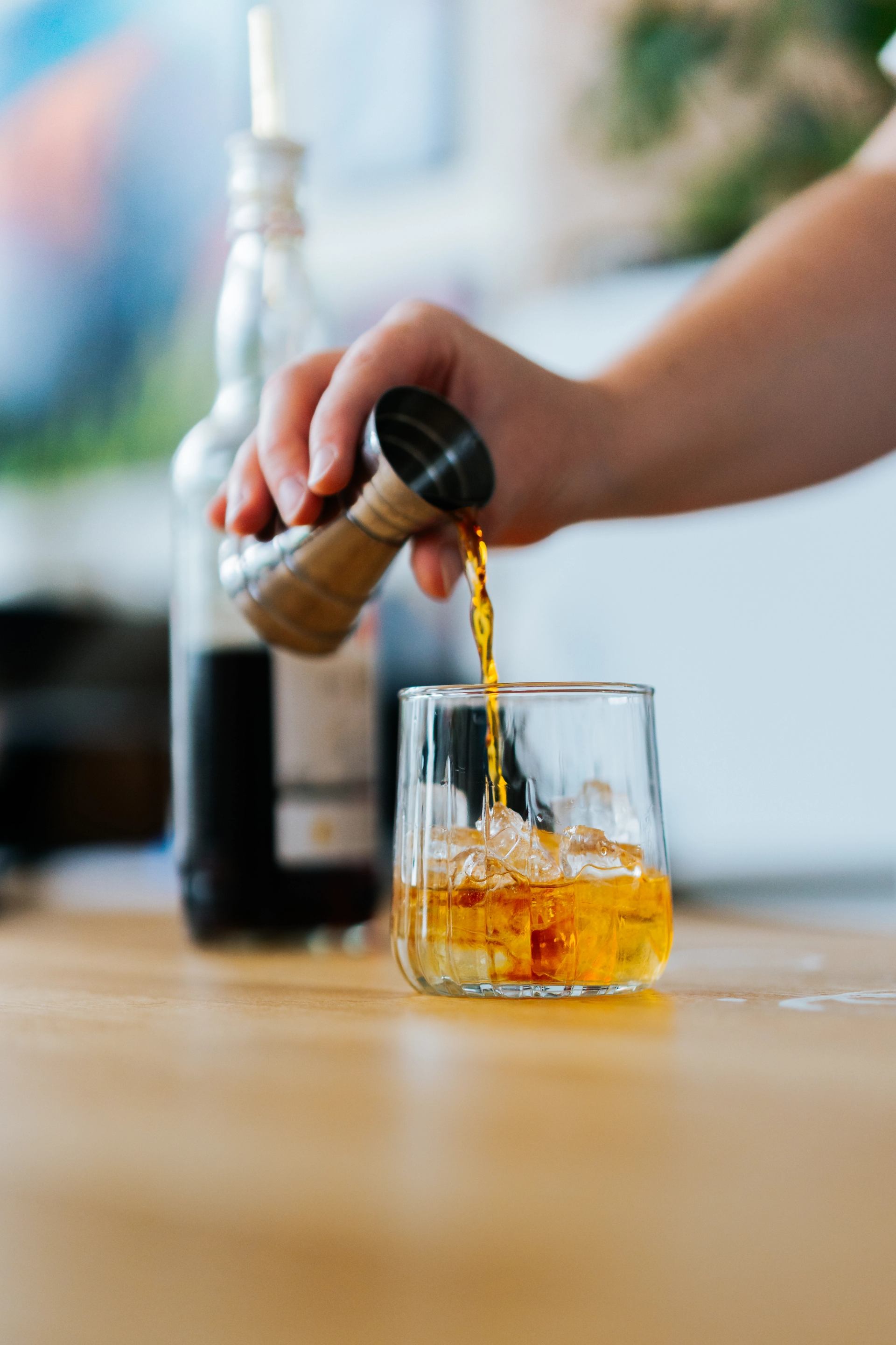 A drink being poured over ice into a short glass on a wooden bar with bottles nearby.