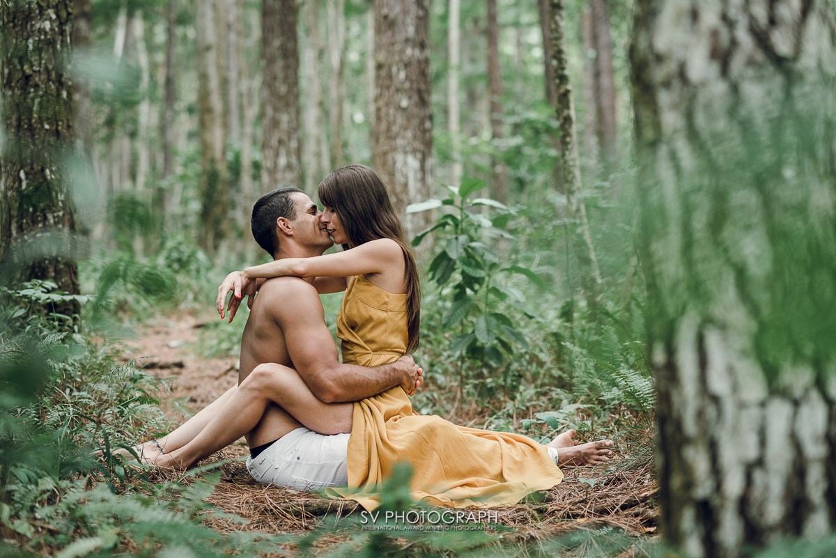 Couple sits closely on forest floor, surrounded by tall trees and lush greenery, sharing a kiss in a peaceful, romantic moment.