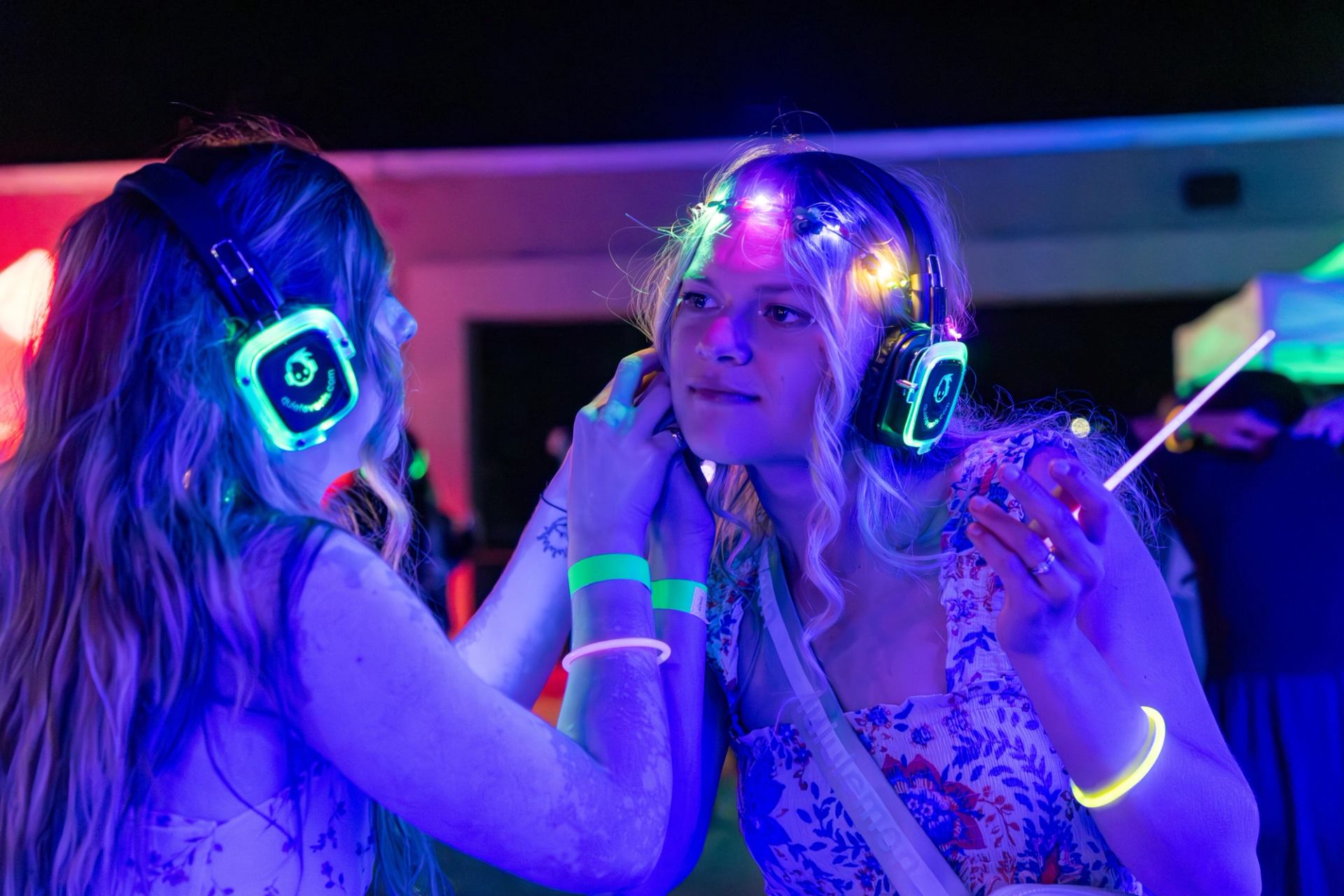 Two people wearing glowing headphones and bracelets stand close under neon party lights.