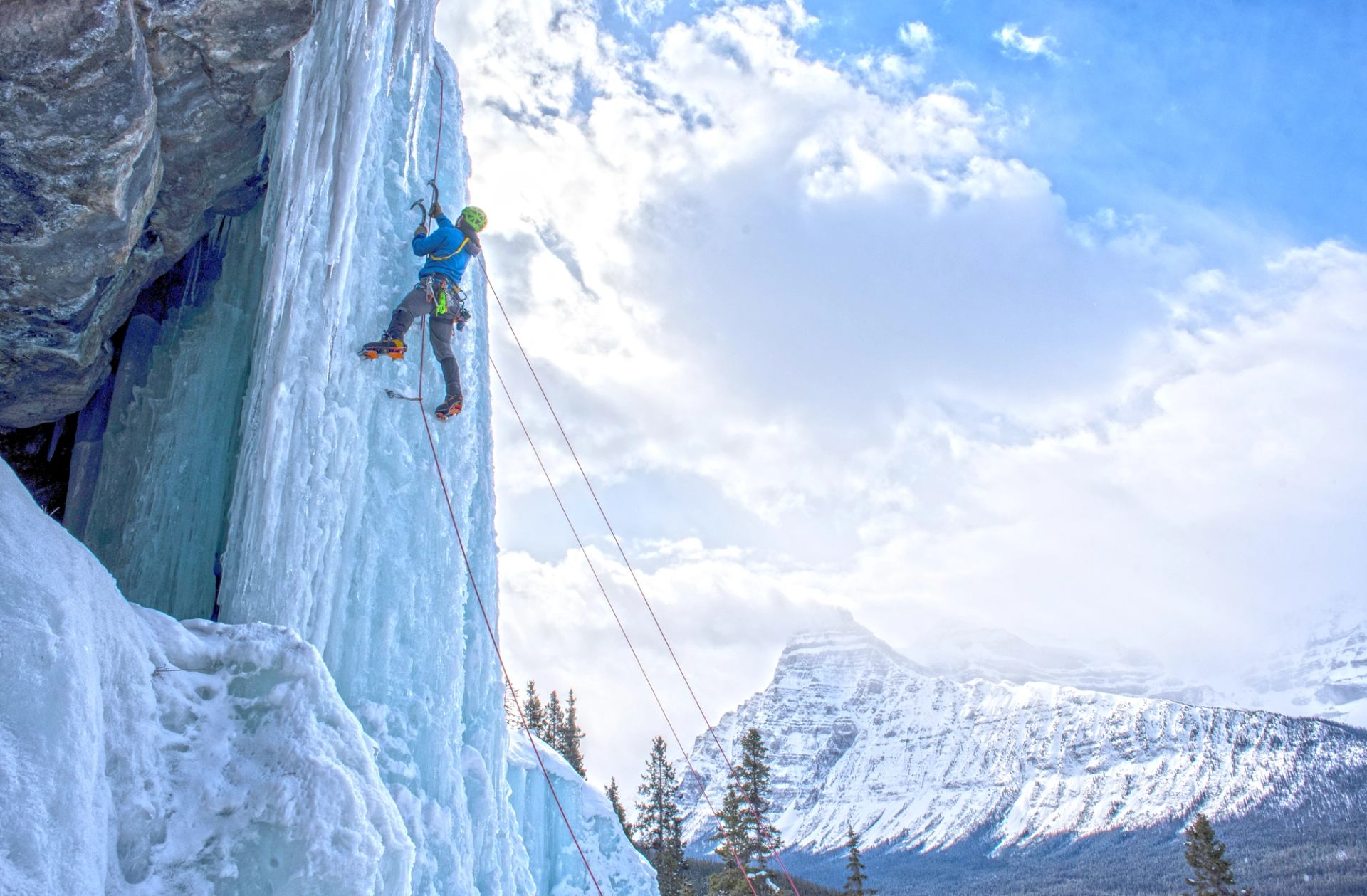 Ice climber ascends frozen waterfall with ropes, snowy mountain range in background.