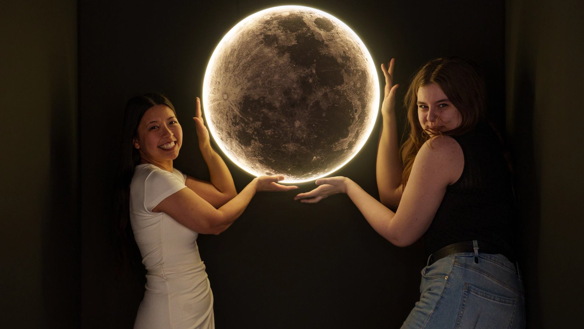 Two people pose beside a glowing full moon wall display in a dark room.