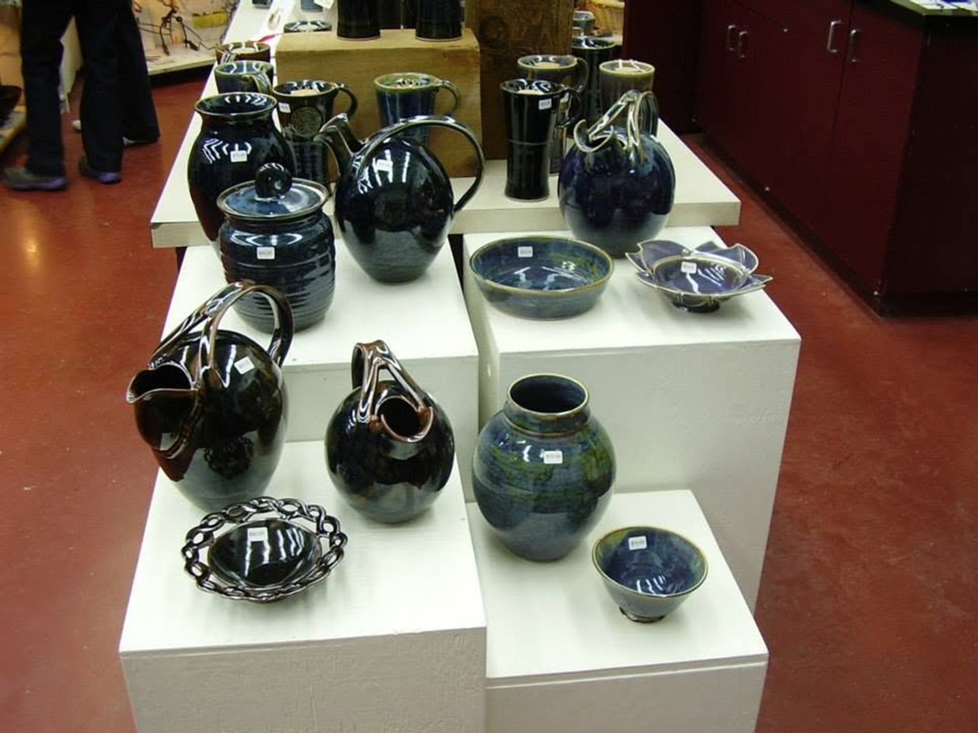 Glazed ceramic bowls, vases, and pitchers are arranged on white display stands inside a pottery stud