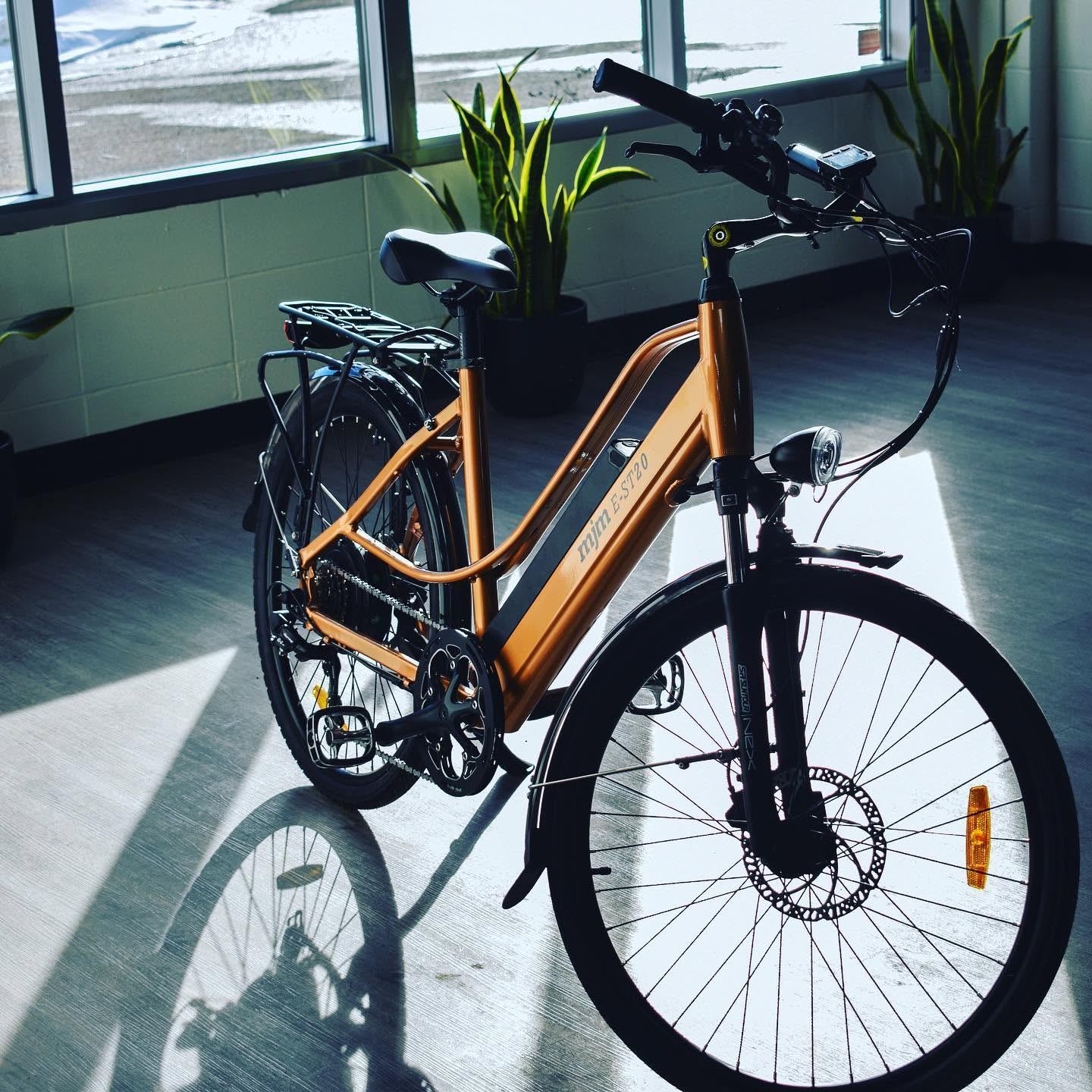 An orange electric bicycle is displayed indoors near large windows, with sunlight casting shadows across the floor and potted plants in the background.