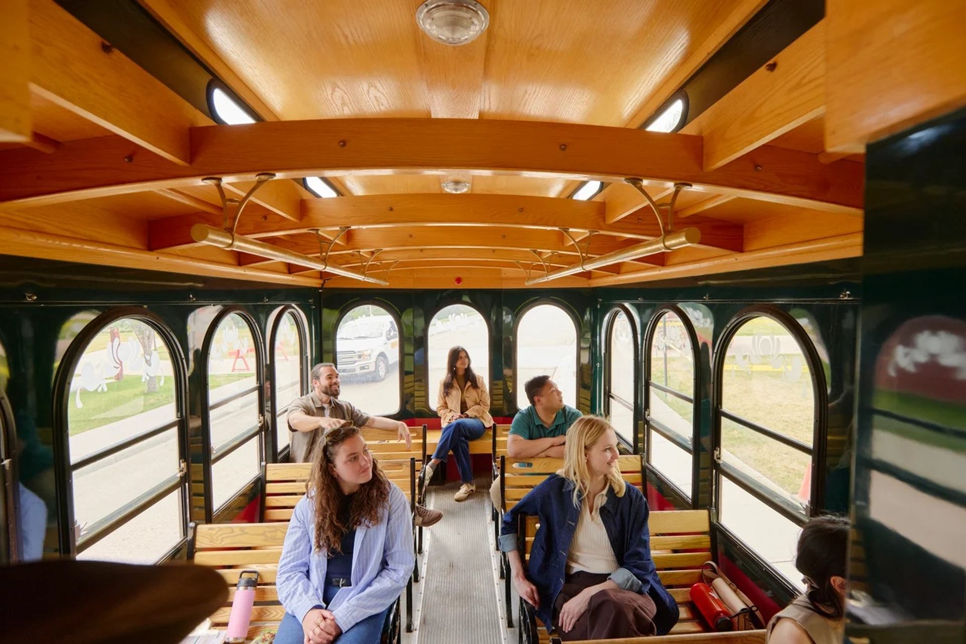 Passengers sit on wooden benches inside a trolley, looking out arched side windows.